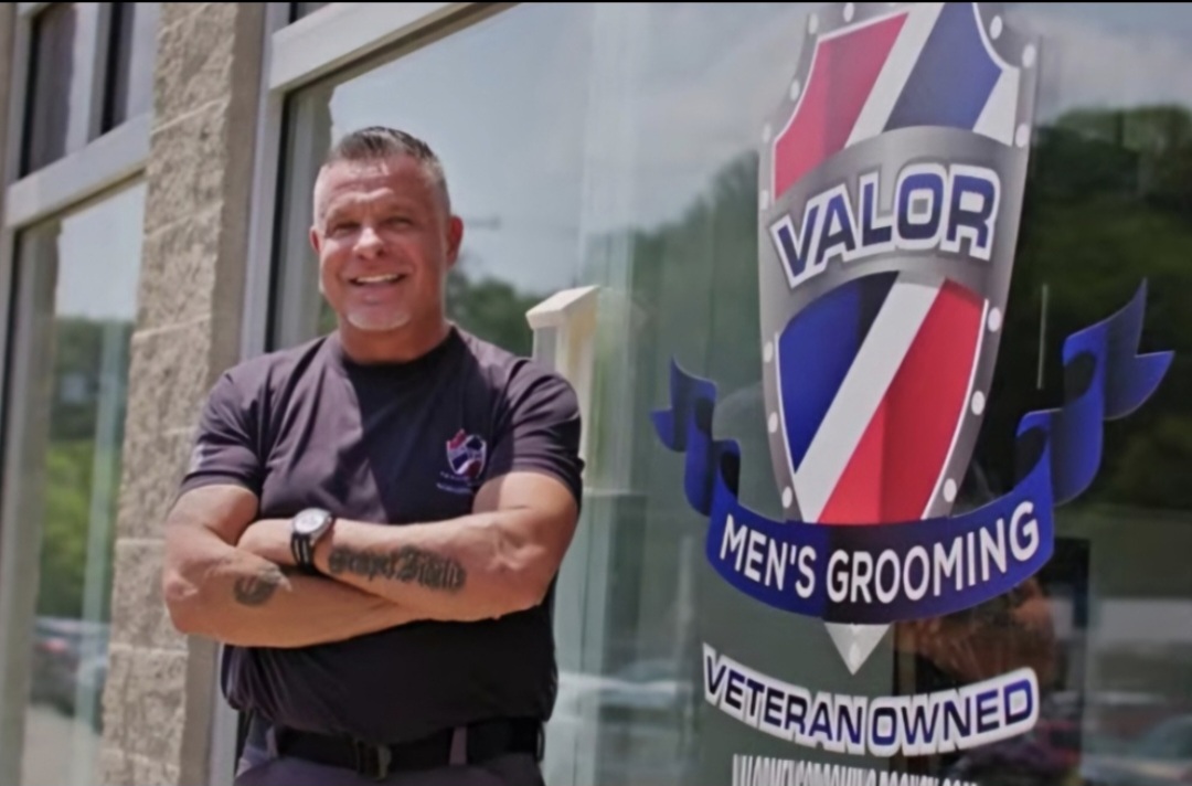 Smiling man standing outside next to a Valor men's grooming sign, arms crossed, wearing a black shirt.