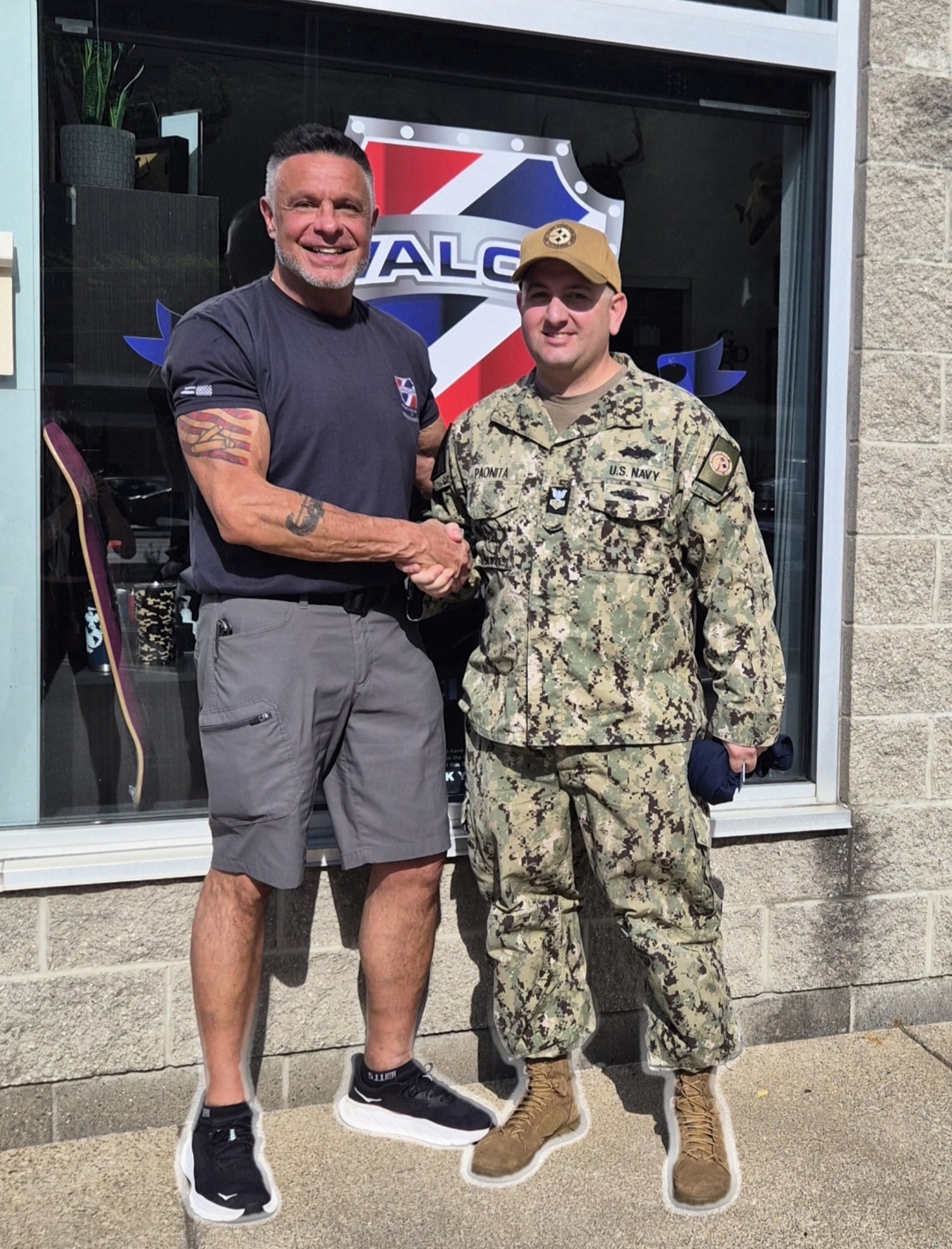 Two men shake hands outside a building, one in casual clothes and the other in military uniform, smiling.