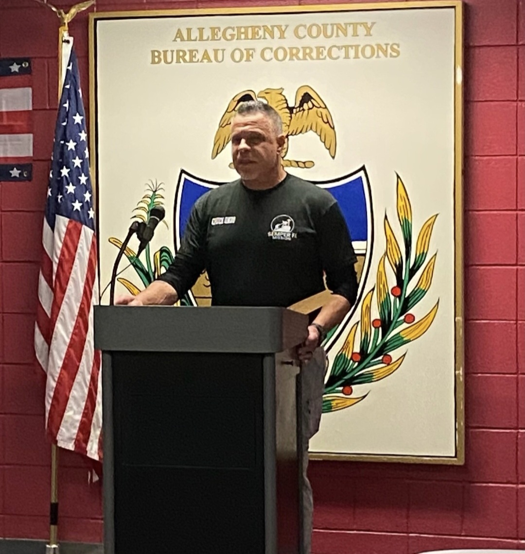 Man standing at a podium in front of a sign for Allegheny County Bureau of Corrections, with an American flag to the left.