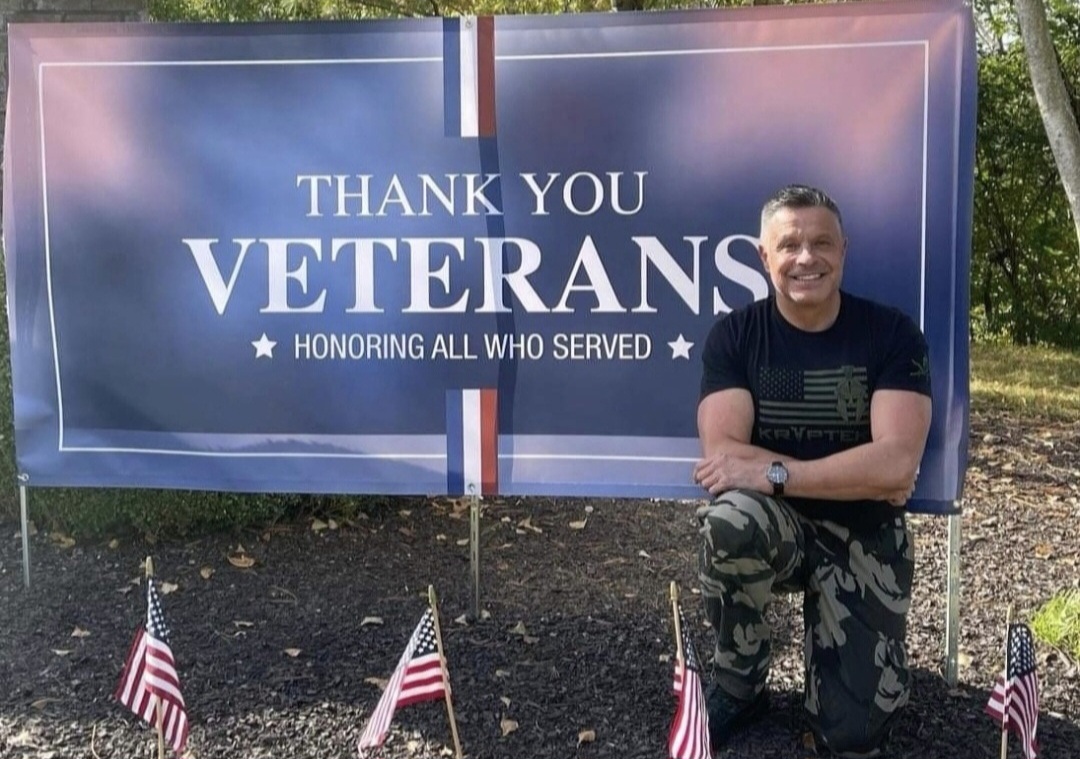 Man standing outdoors next to a large sign that reads 'Thank You Veterans, Honoring All Who Served' with small American flags at the base.