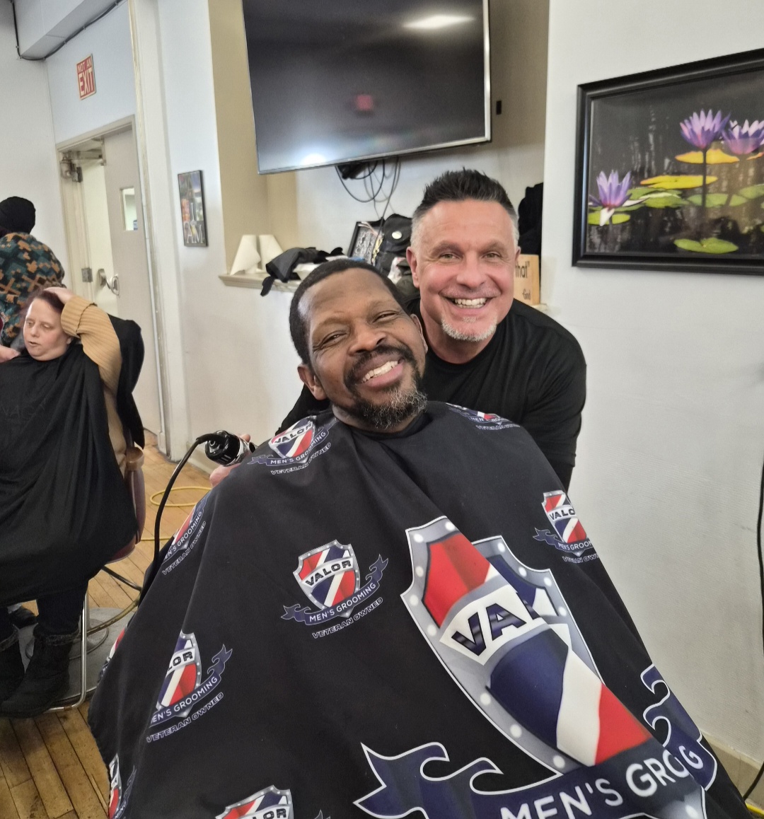 Two men smiling, one with a beard, in a barbershop chair with a cape, in a room with a TV and artwork.