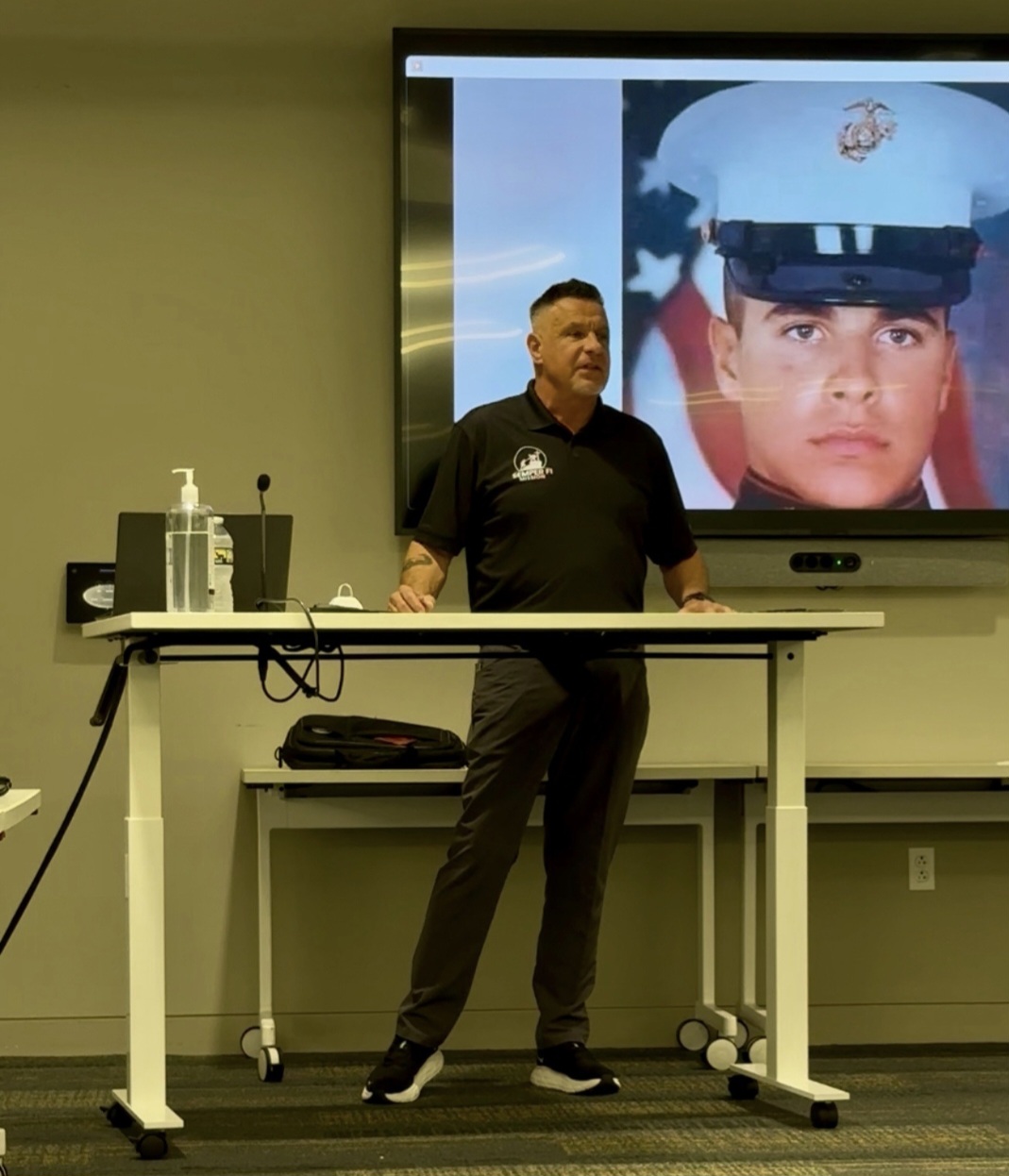 Man standing in front of a presentation screen with a photo of a woman in a military uniform, in a classroom or conference room.