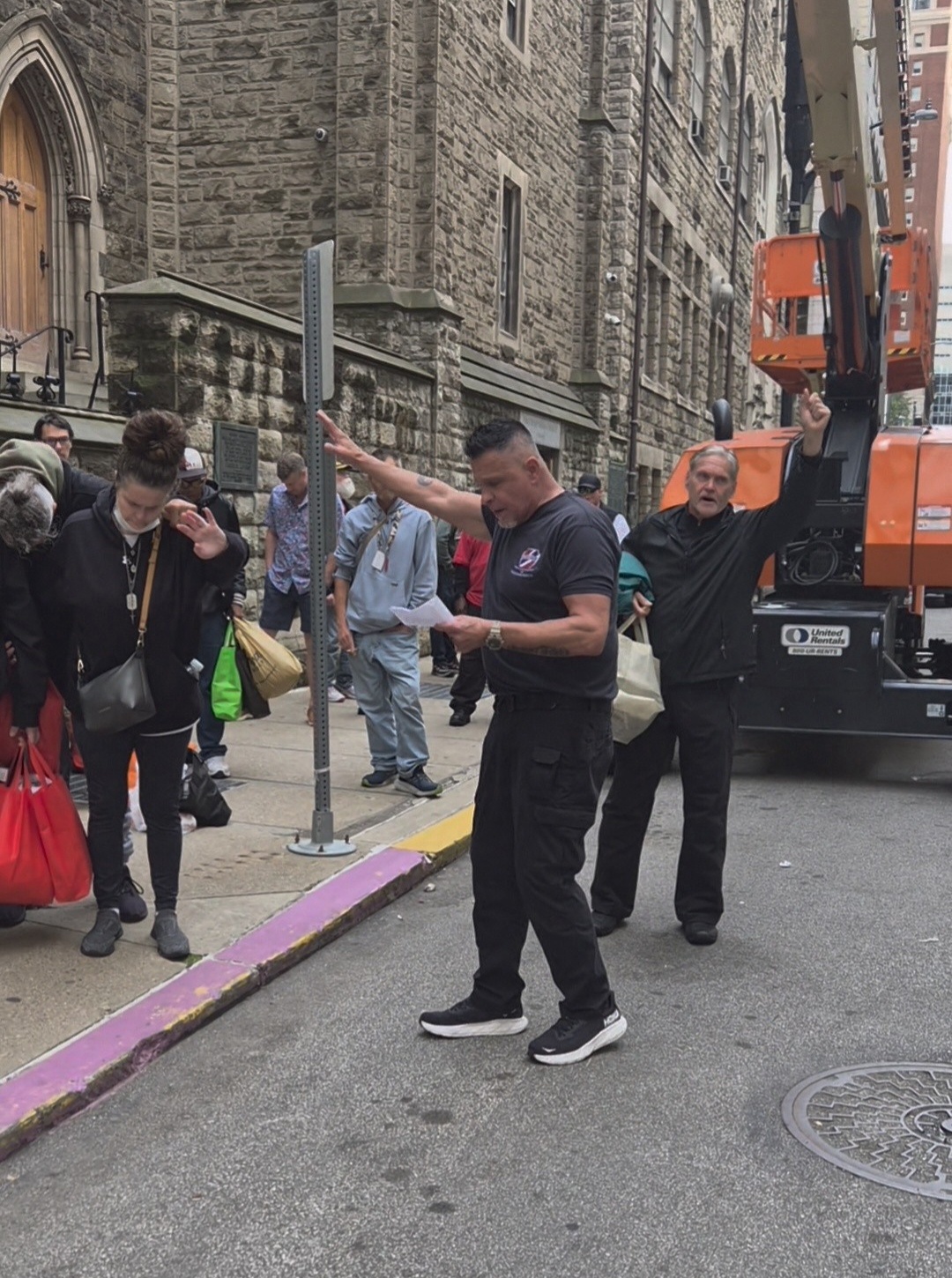 Group of people standing on a city street near a stone building, with a person looking at a phone in the center.
