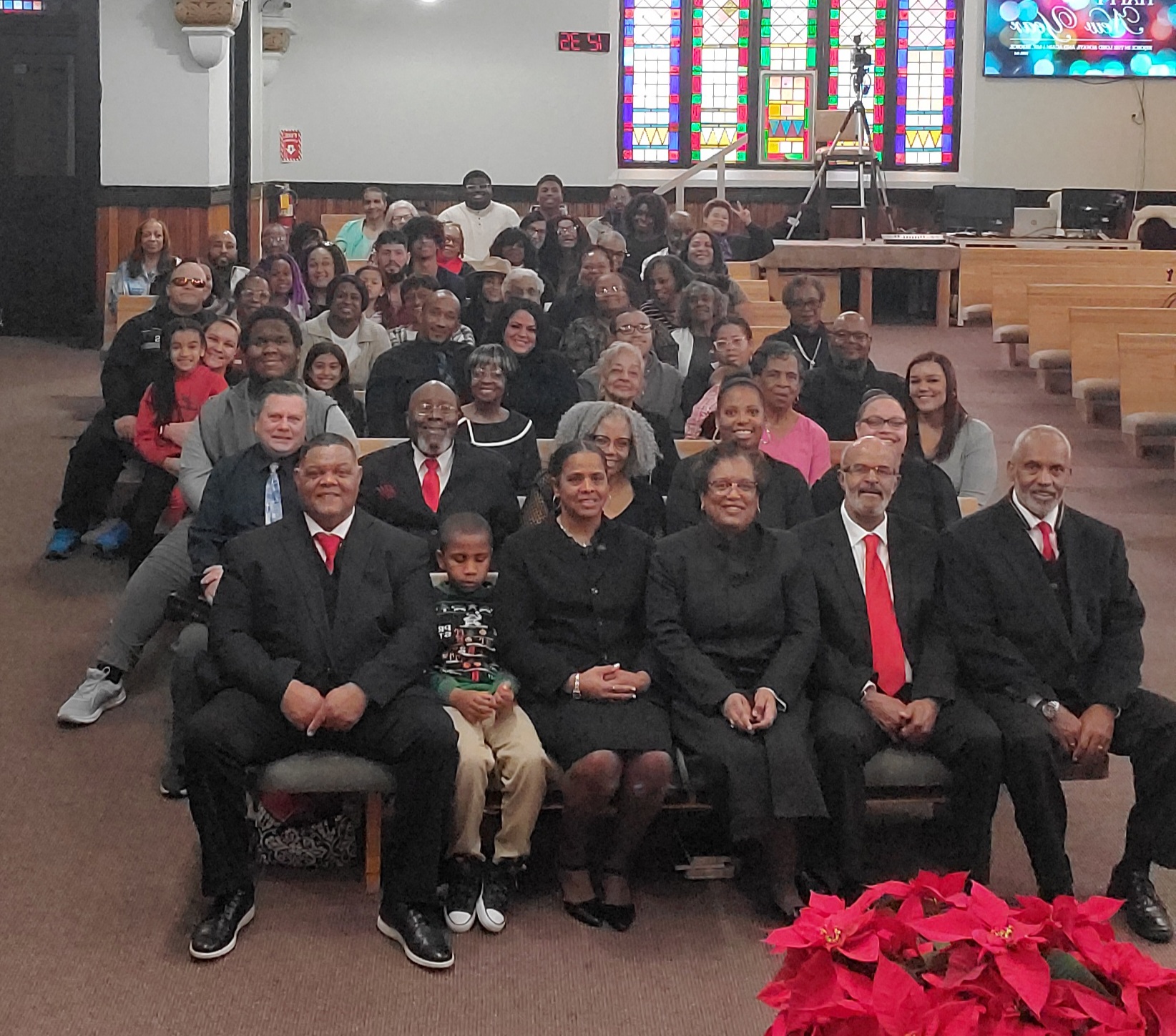 Group of people seated in a church, with stained glass windows in the background, some wearing formal attire, and a poinsettia plant in the foreground.