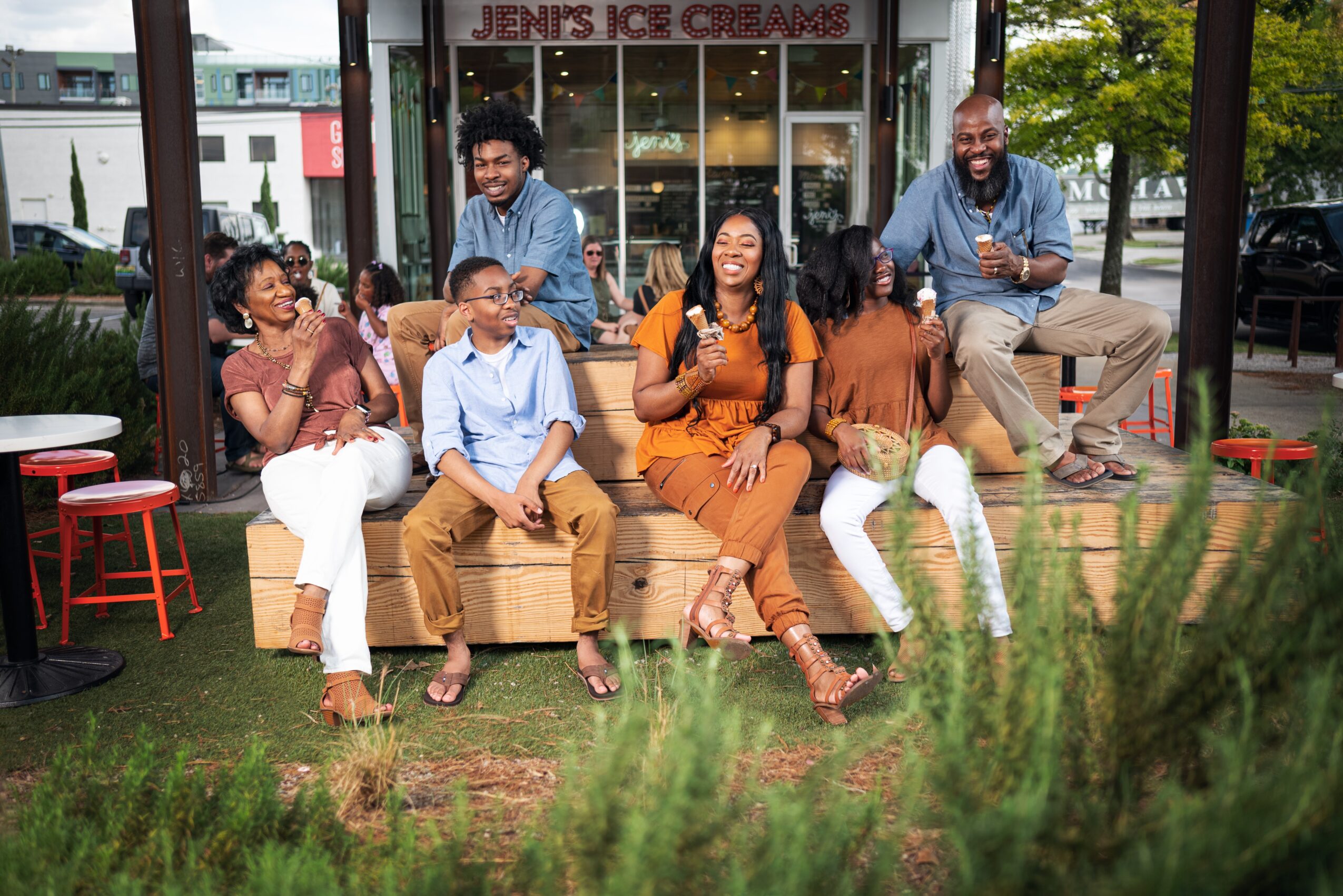 Group of seven people sitting and standing outdoors in front of an ice cream shop, smiling and enjoying each other's company.