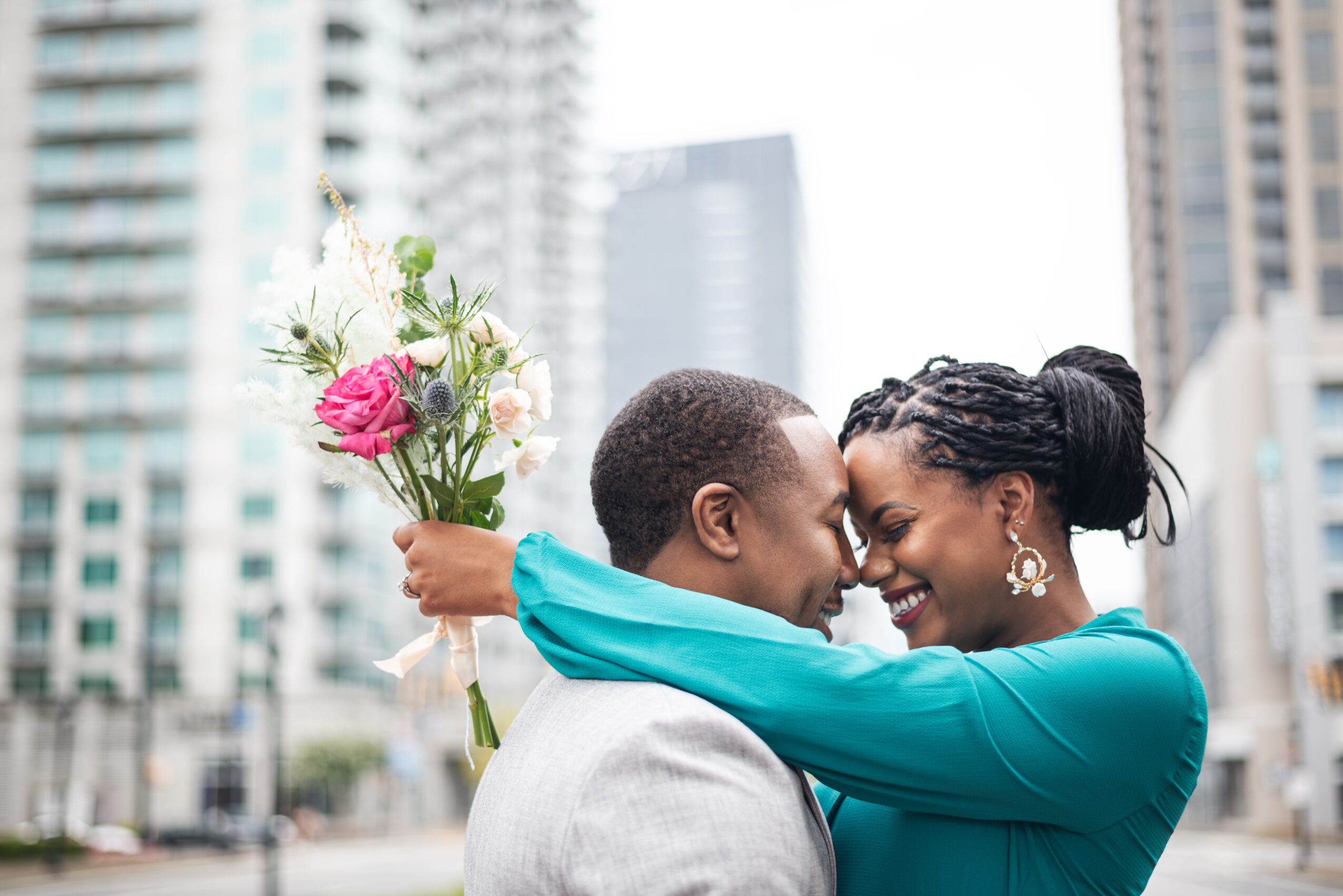 Two women face each other closely, one holding a bouquet of flowers, in an urban setting with tall buildings behind them.