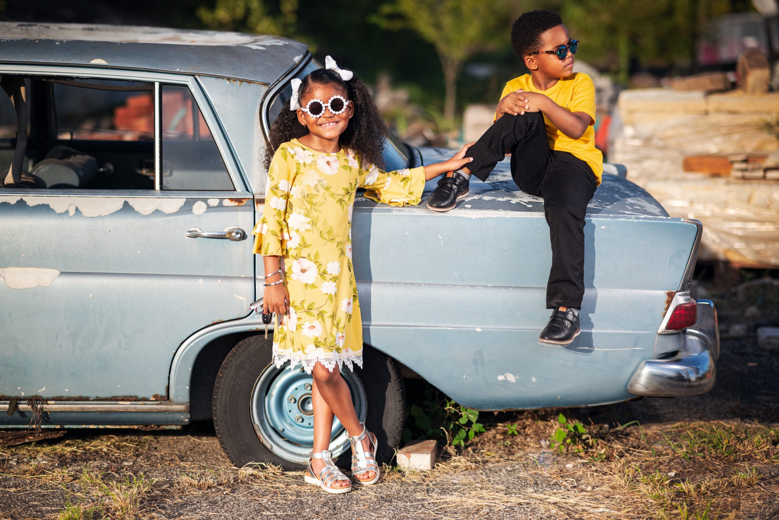 Two children playing near a vintage car outdoors, girl in a yellow dress and sunglasses, boy in a yellow shirt sitting on the car's trunk.