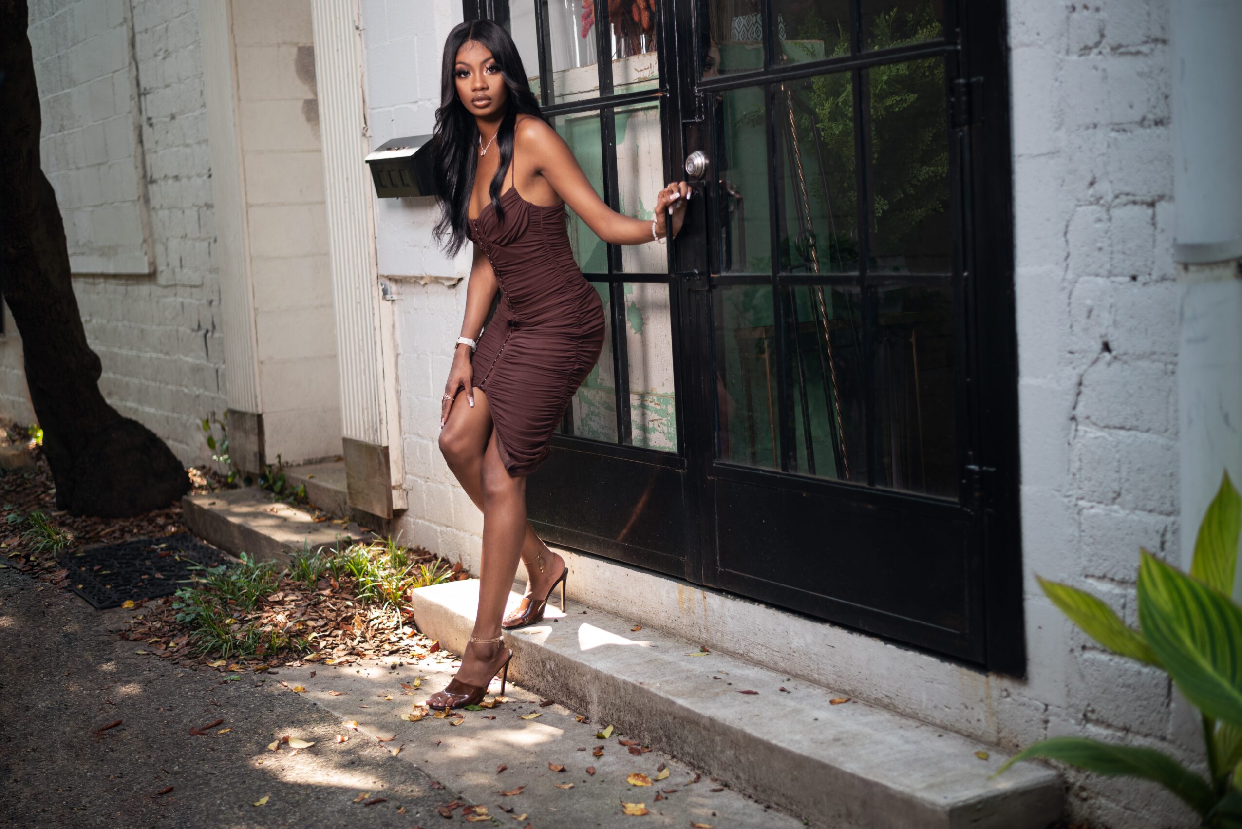 Woman in a fitted dress standing outside near a black door, holding a handbag, with a white brick wall behind her.