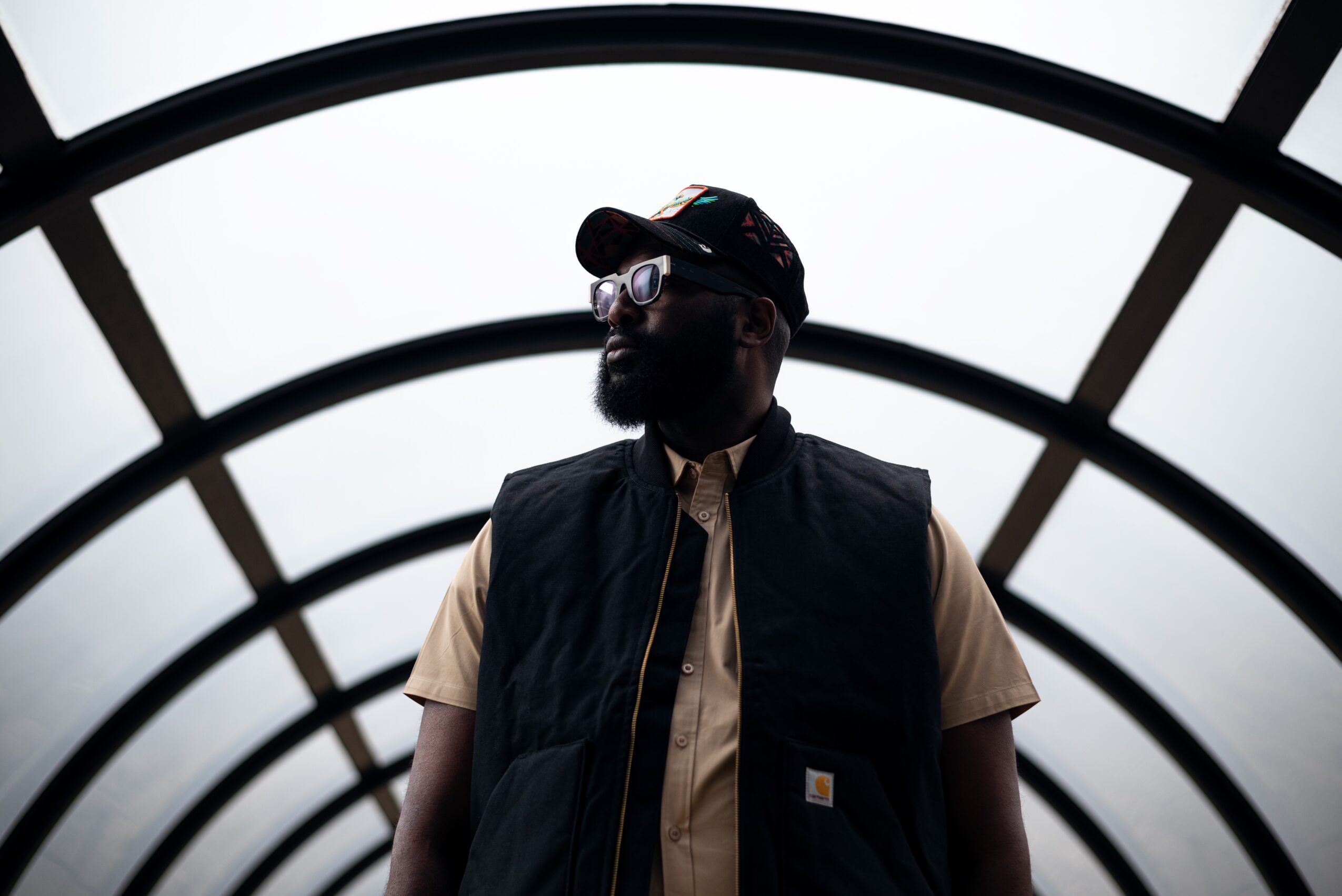 Person wearing glasses, cap, and vest standing in a tunnel with curved ceiling, backlit by bright light.