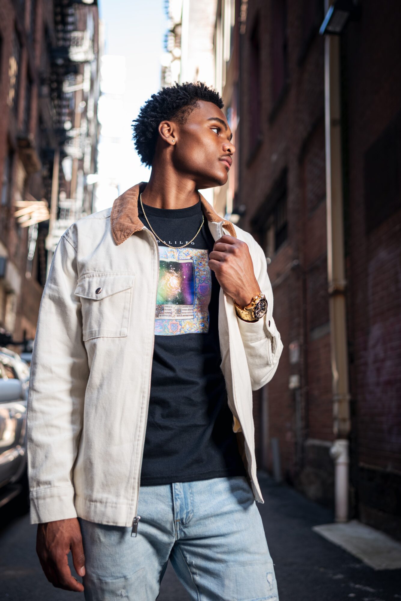 Young person with short curly hair standing on city street, looking to the side, wearing a beige jacket and black graphic t-shirt.