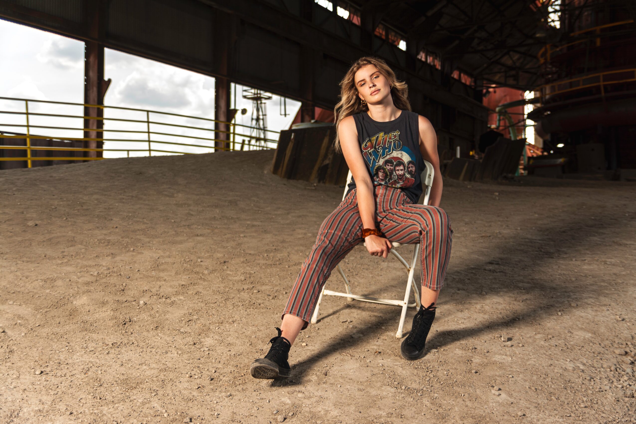 Young woman with wavy hair sitting on a chair in an industrial space, wearing a graphic tank top and striped pants.