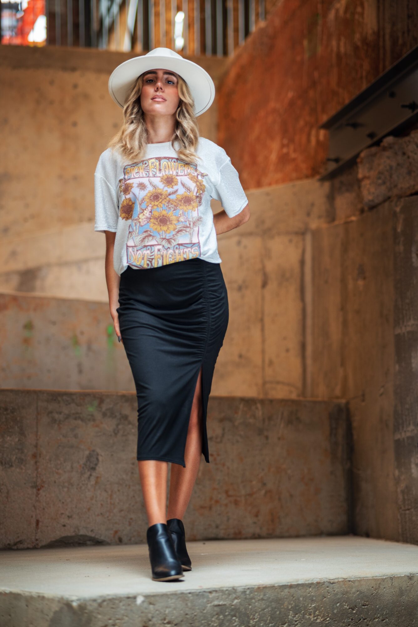 Woman in a white hat, patterned top, and black skirt walking on a stage with wooden background.