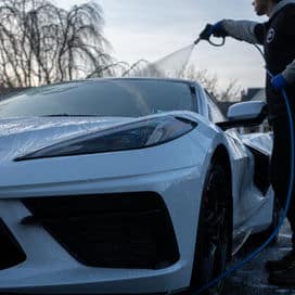 Person washing a white sports car outdoors with trees in the background.