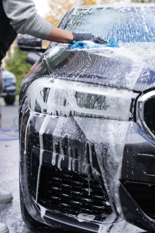 Person washing a black car with soap and water outdoors, hand wearing a black glove, trees in background.