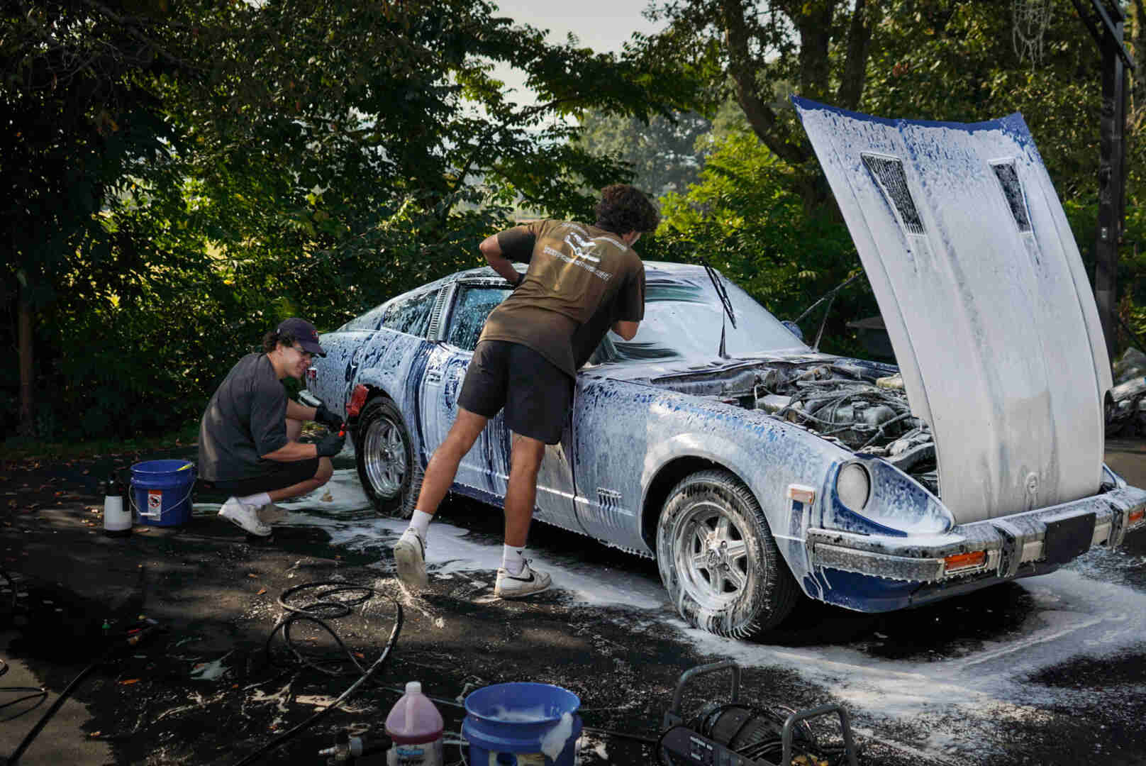 Two people wash a blue car outdoors with trees in the background, one scrubbing the side and the other working near the hood.