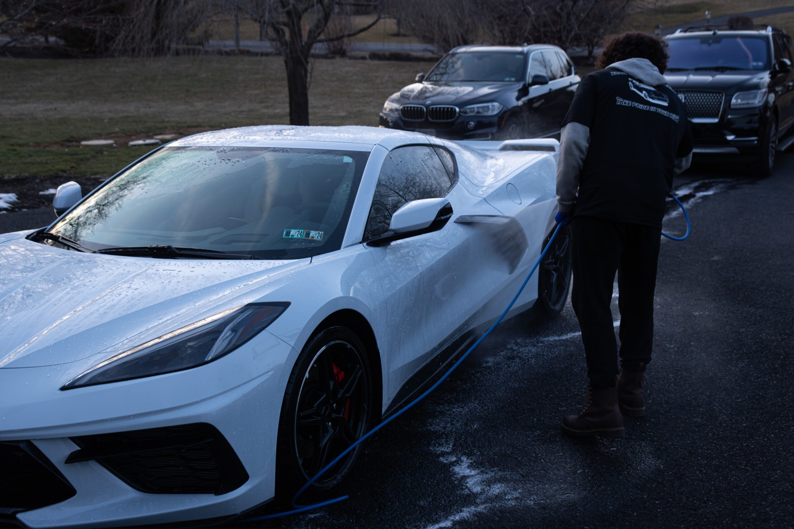 Person washing a white sports car outdoors with other vehicles in the background.