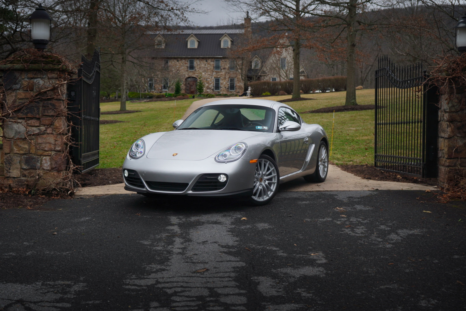 Silver sports car parked between stone gateposts with open gates, trees, and a building in the background.