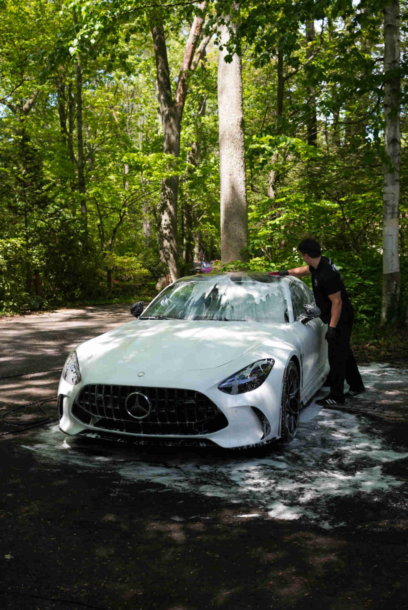 Person washing a white sports car in a forested area with trees and sunlight, water on the ground.