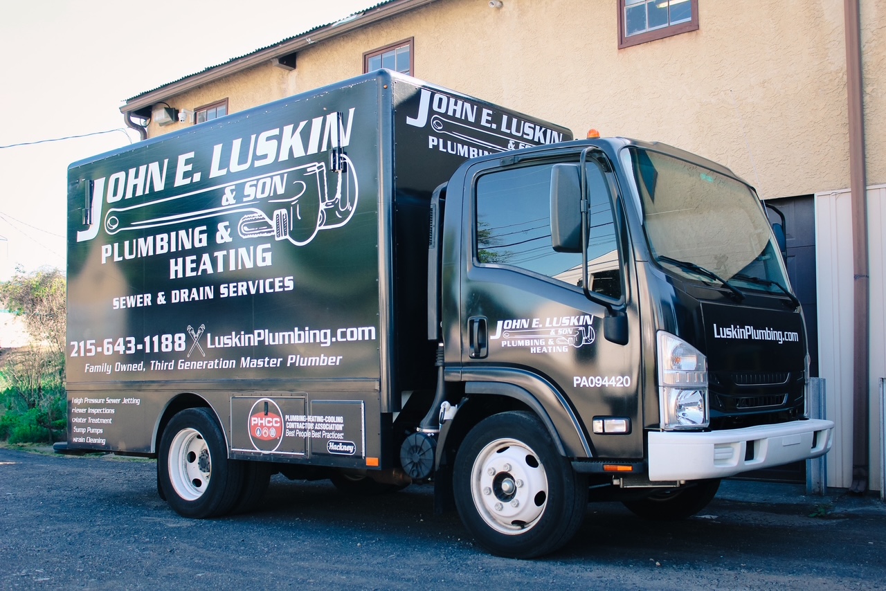 Black plumbing and heating truck parked on street with building in background, displaying company name and contact information.