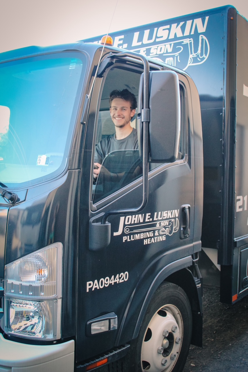 Young man smiling inside a black plumbing service truck with company logo and contact information, parked outdoors.