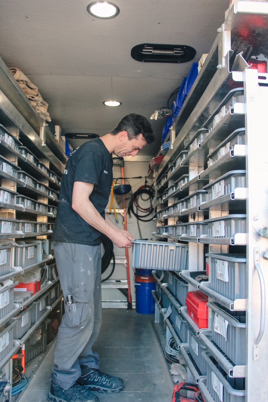Person in a storage room with shelves filled with containers and tools, holding a small object, facing right.