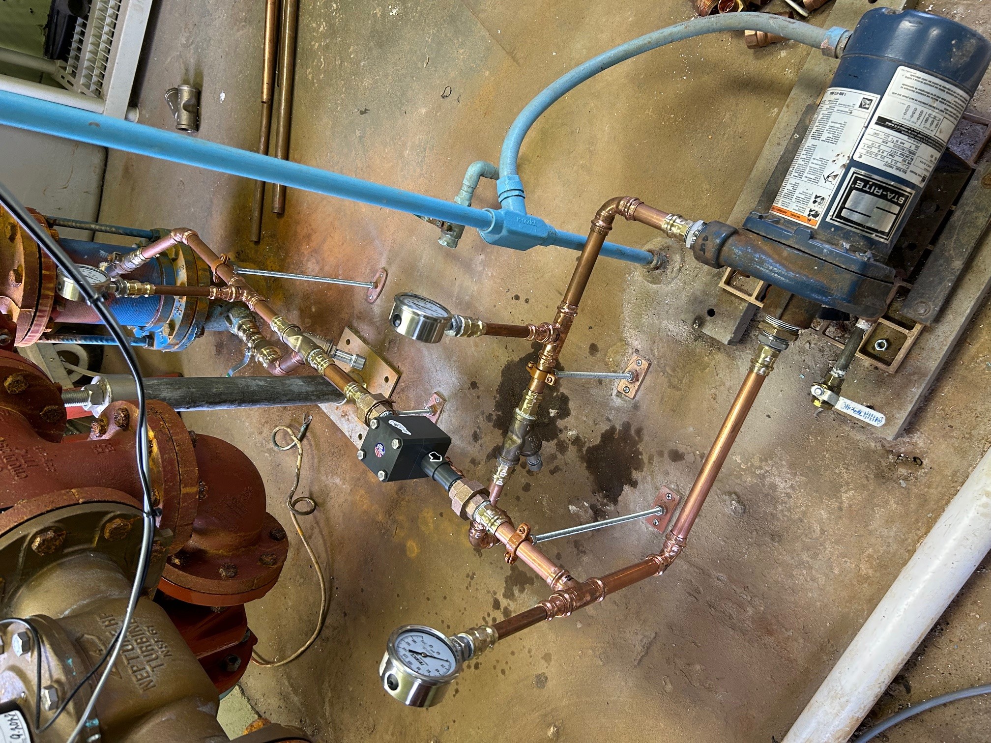 Pipes and a compressor mounted on a wall with water and rust stains, connected by blue and copper pipes.