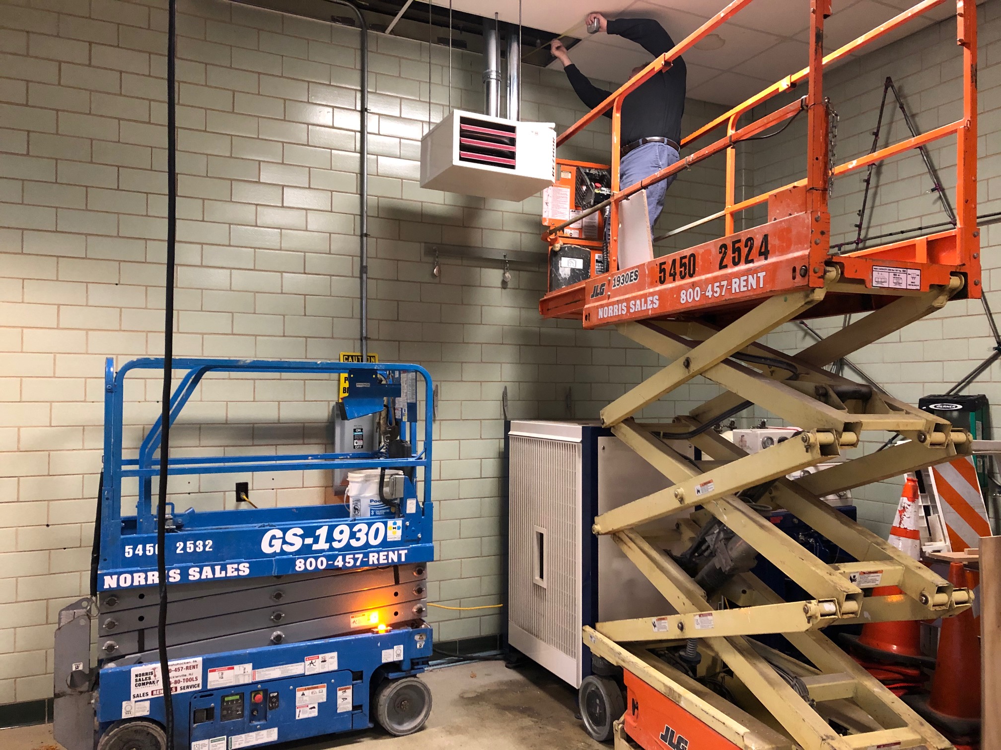 Person on orange scissor lift working near ceiling vent in industrial space with brick wall.