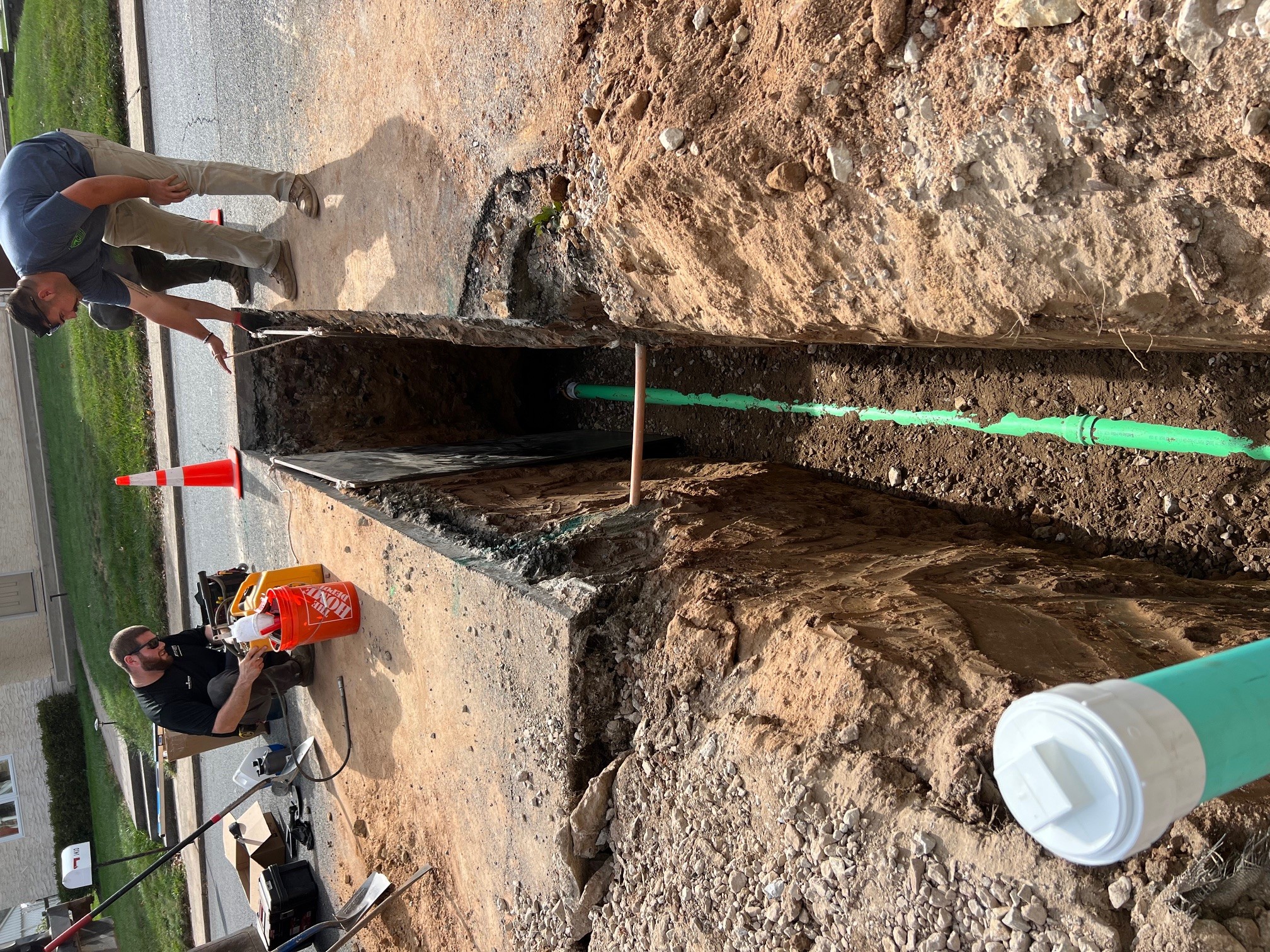 Two workers at a construction site with a long trench, pipes, and tools, on a sunny day. One worker is standing, the other is crouching.