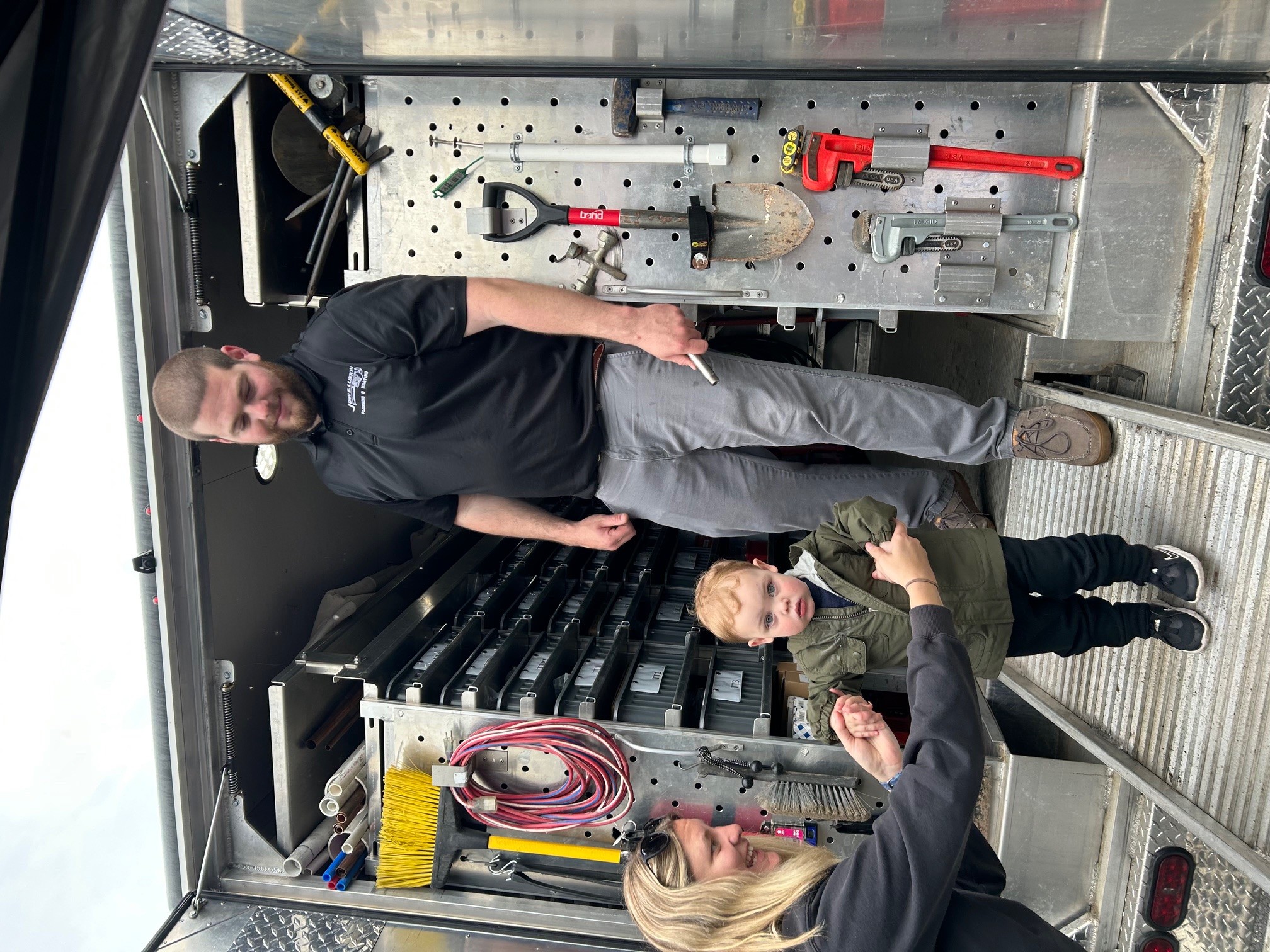 Two people inside a service truck, one sitting and one standing, with tools and equipment on the wall behind them.