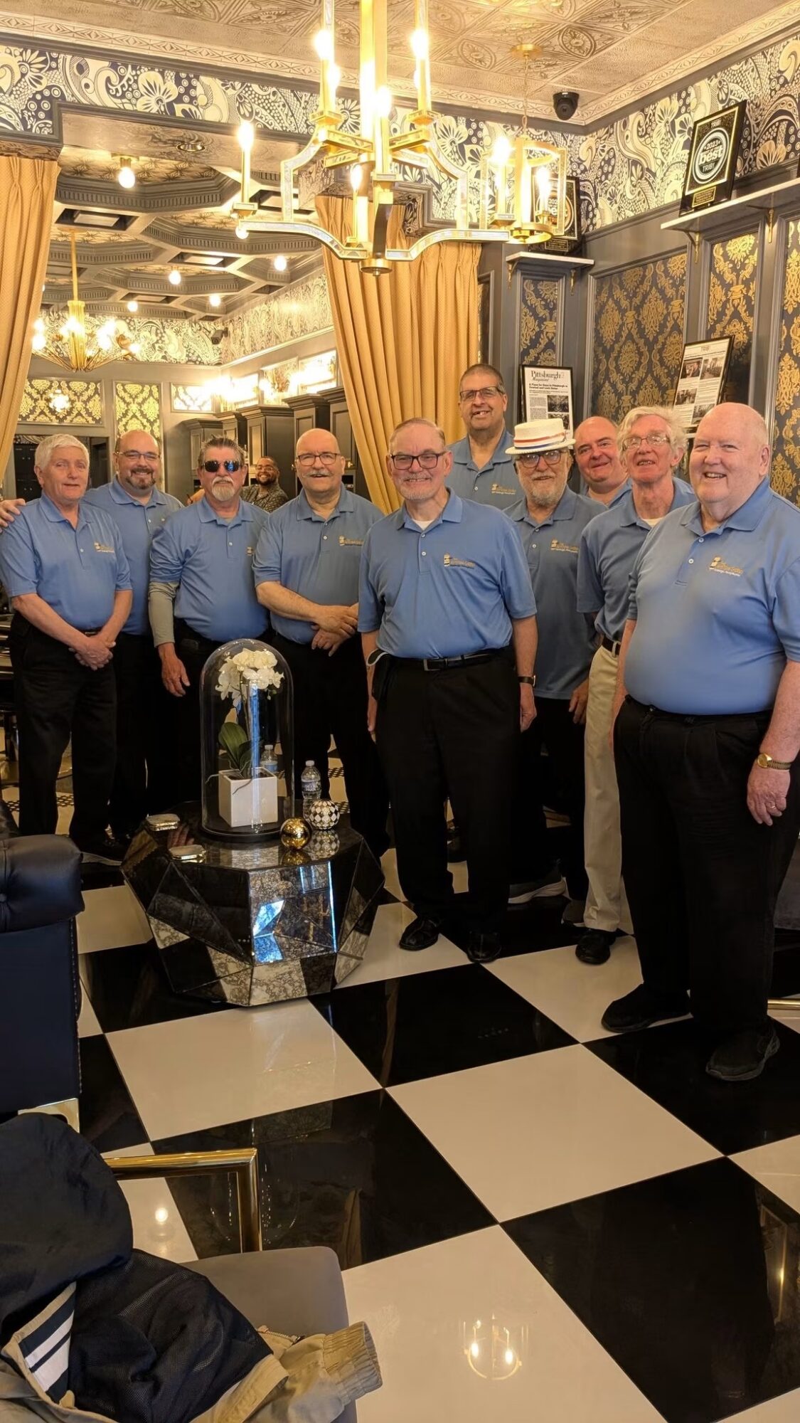 Group of nine men in matching blue shirts standing in a decorated room with black and white checkered floor, chandelier, and ornate walls.