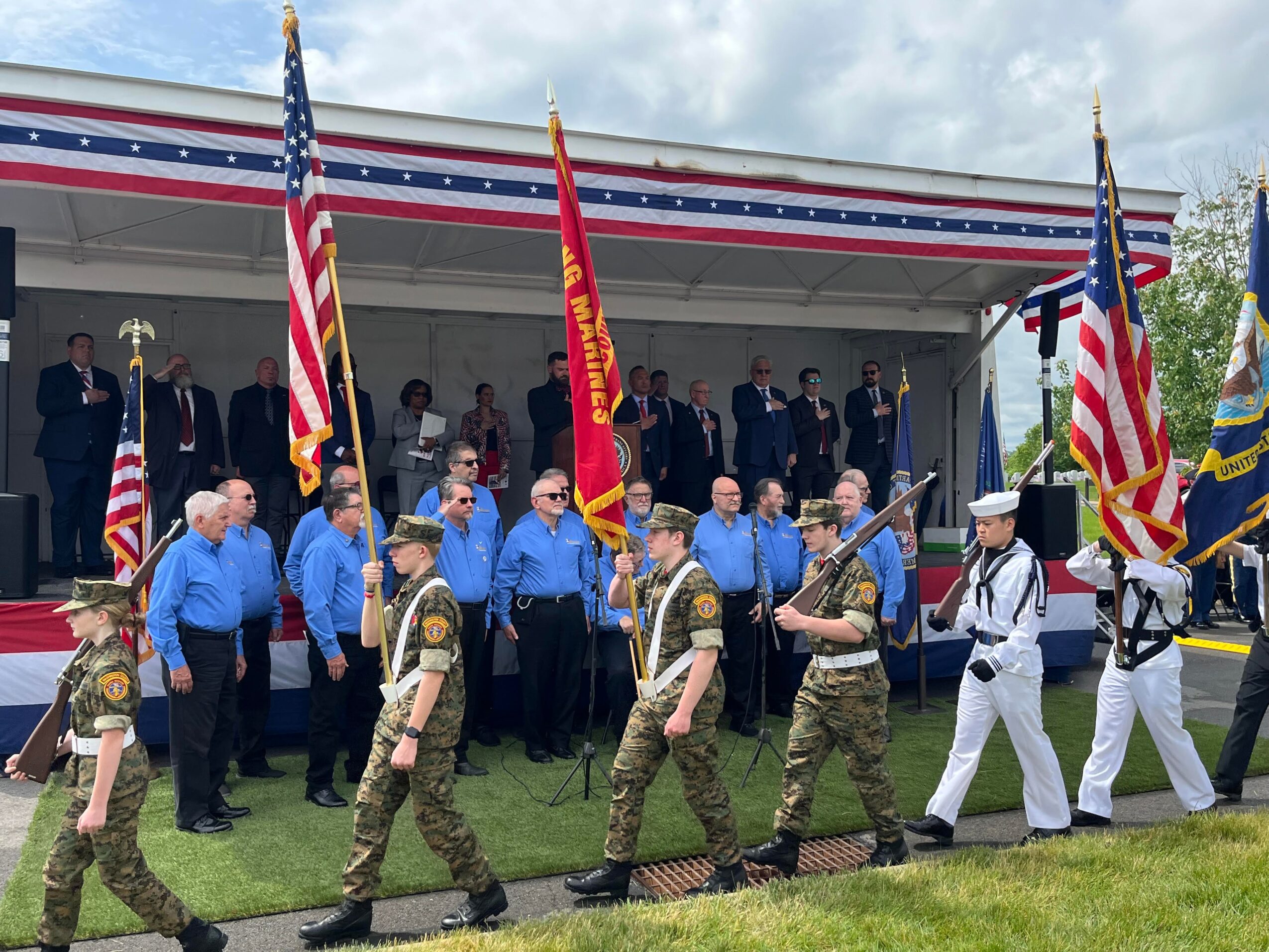Group of people, including military personnel and veterans, standing on a stage with American flags and a red flag, outdoors.