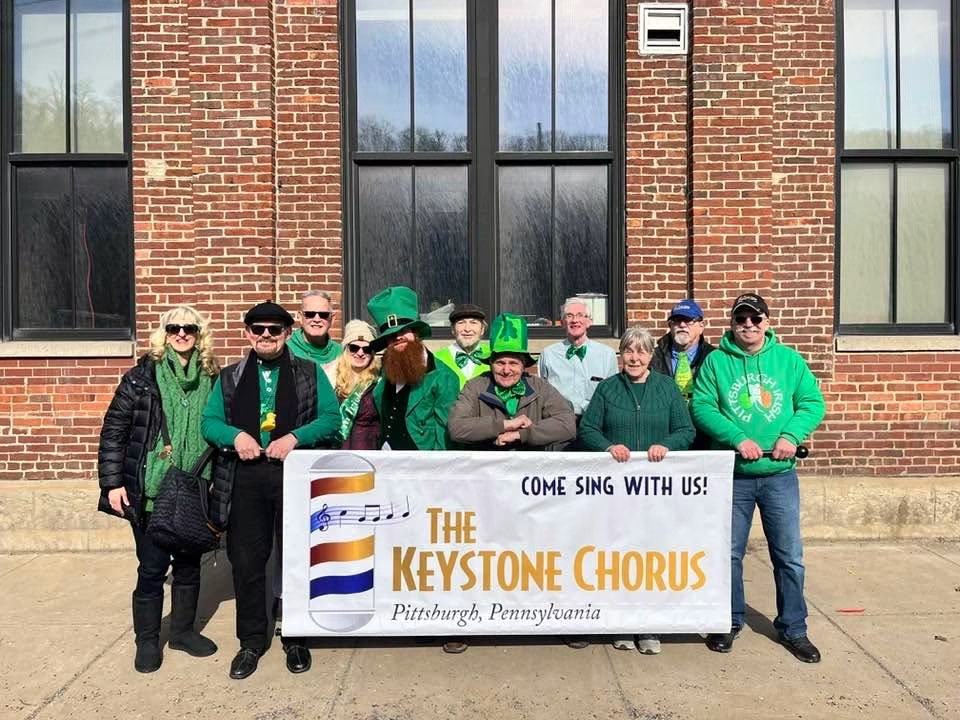 Group of people holding a banner for The Keystone Chorus in front of a brick building with large windows, sunny day.