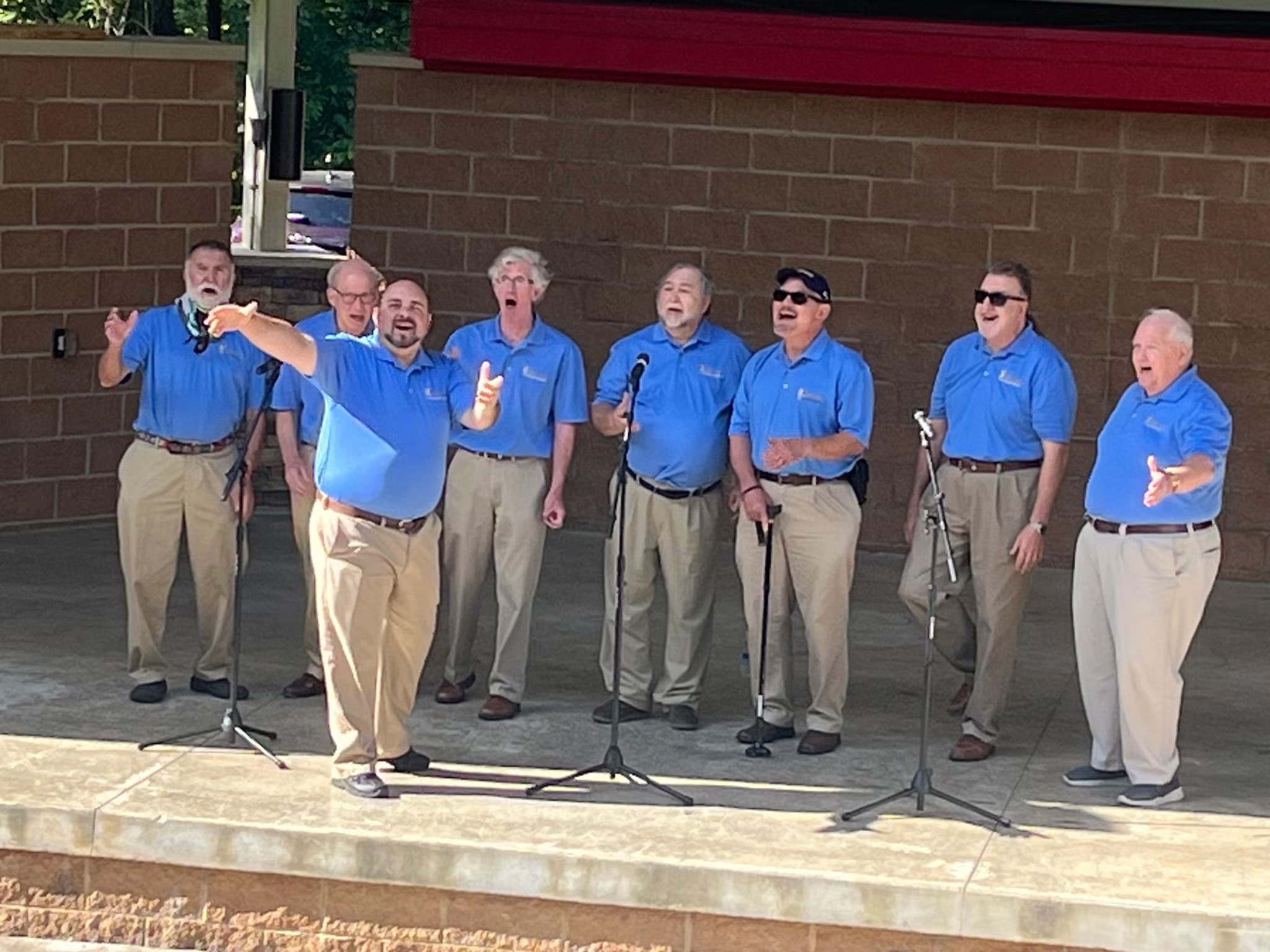 Group of eight men singing outdoors, wearing blue shirts and khaki pants, standing on a stage with microphones.