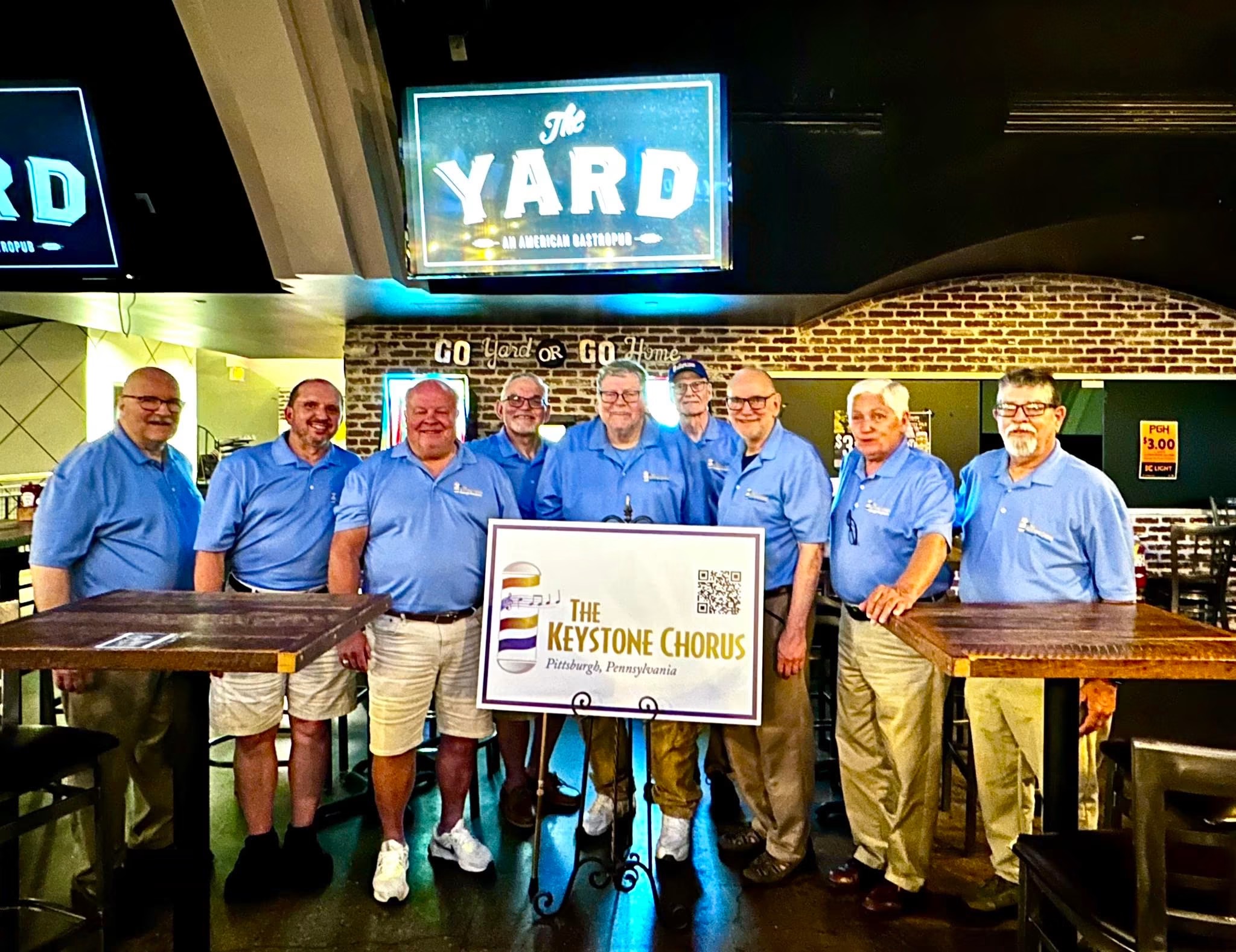 Group of nine men in blue shirts holding a sign for Keystone Chorus, standing inside a venue with signs reading 'The Yard'.