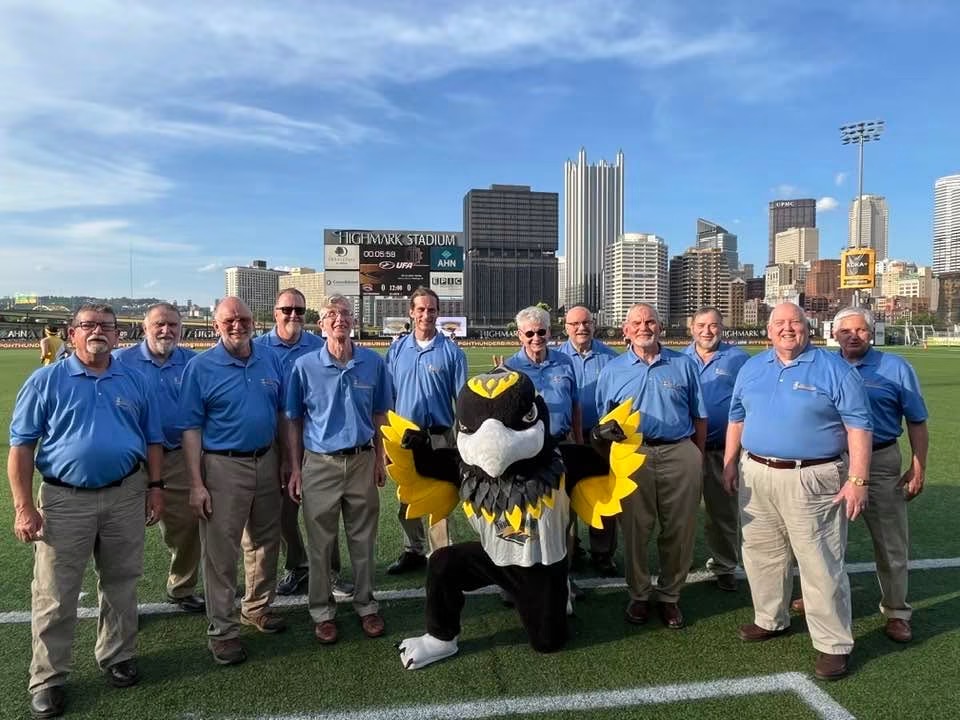 Group of nine people in blue shirts standing on a sports field with city skyline in background.