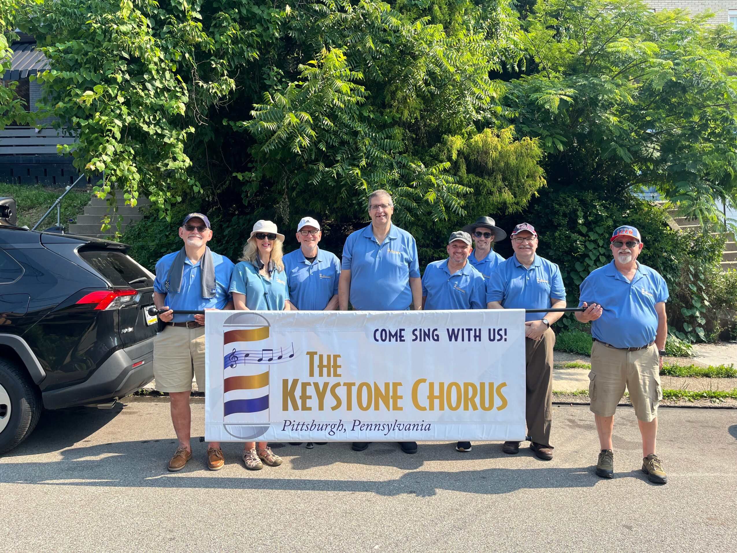Group of seven people holding a banner for The Keystone Chorus outdoors with trees in the background.