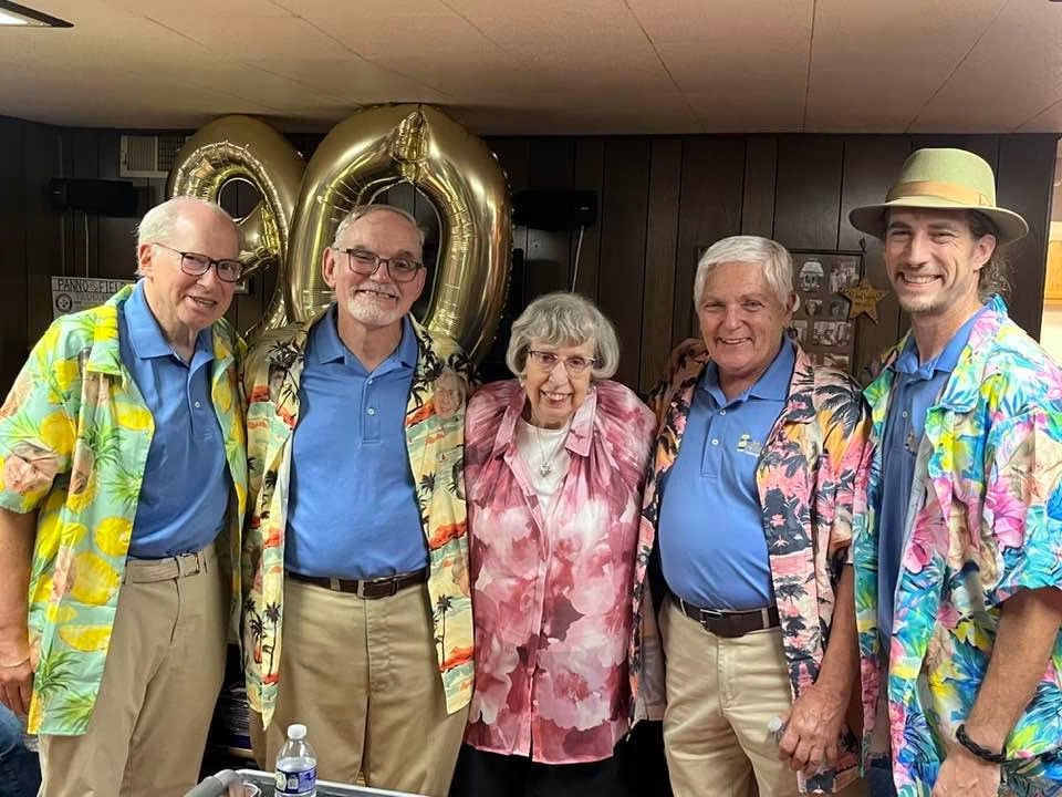 Five people standing together, smiling, with large gold balloons in the background, celebrating a 50th anniversary, indoors.