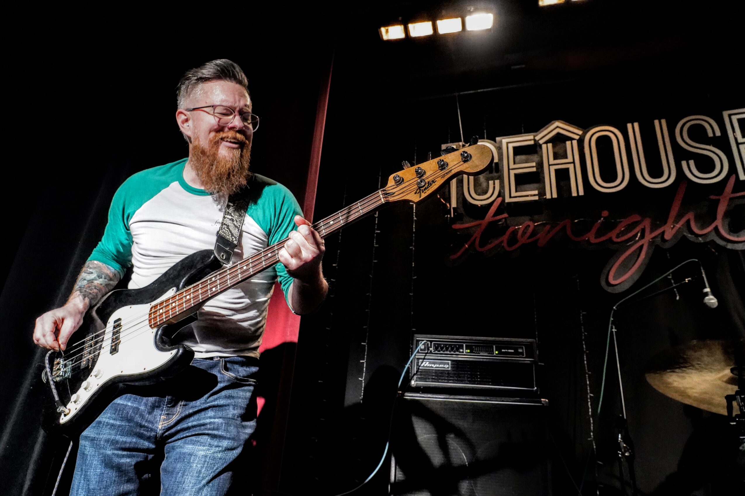 Person with glasses and a beard playing a bass guitar on stage, smiling, with a sign reading 'THE HOUSE Tonight' in the background.