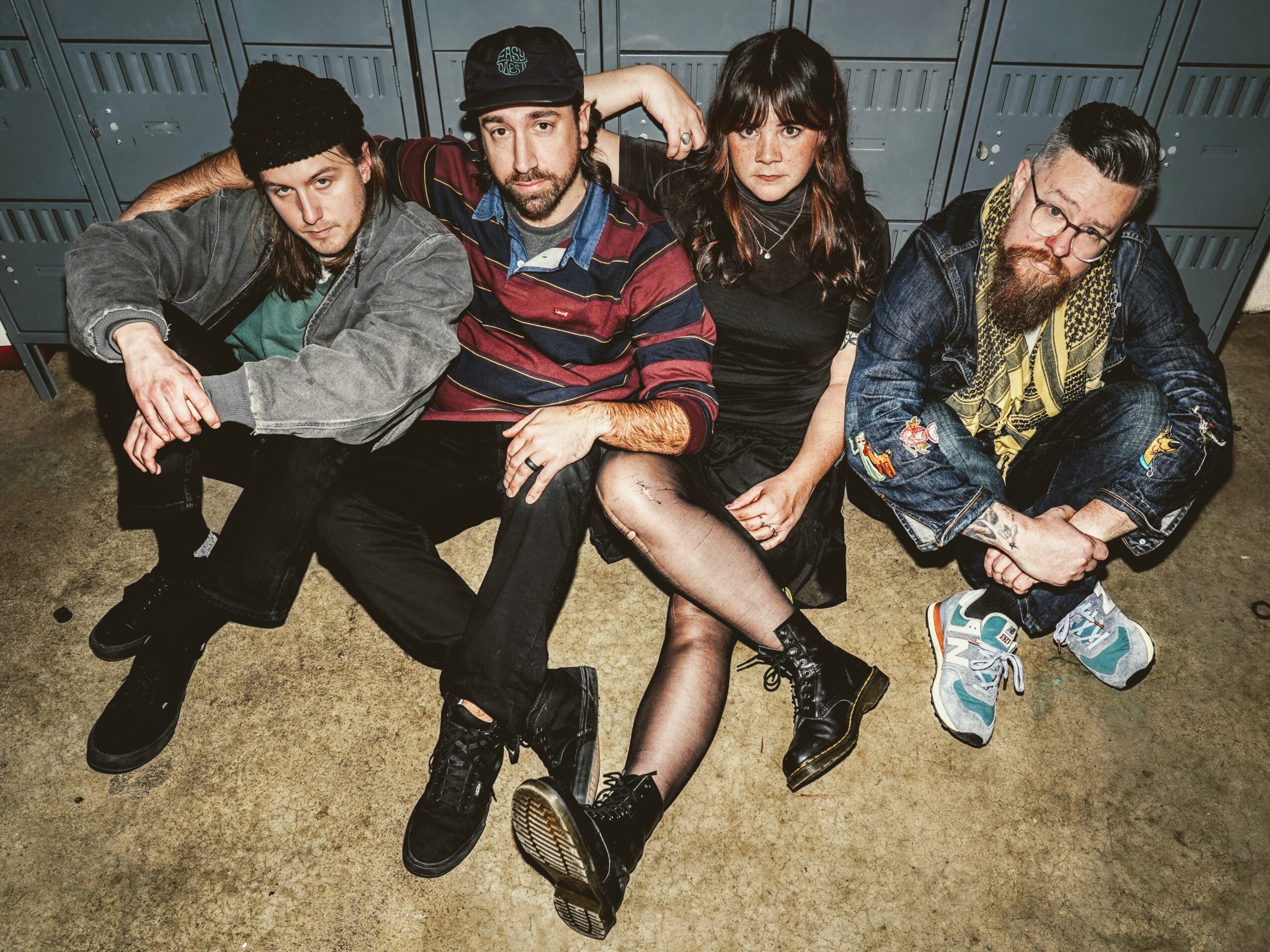 Four people sitting on a bench in front of lockers, posing for the photo, with casual clothing and accessories.