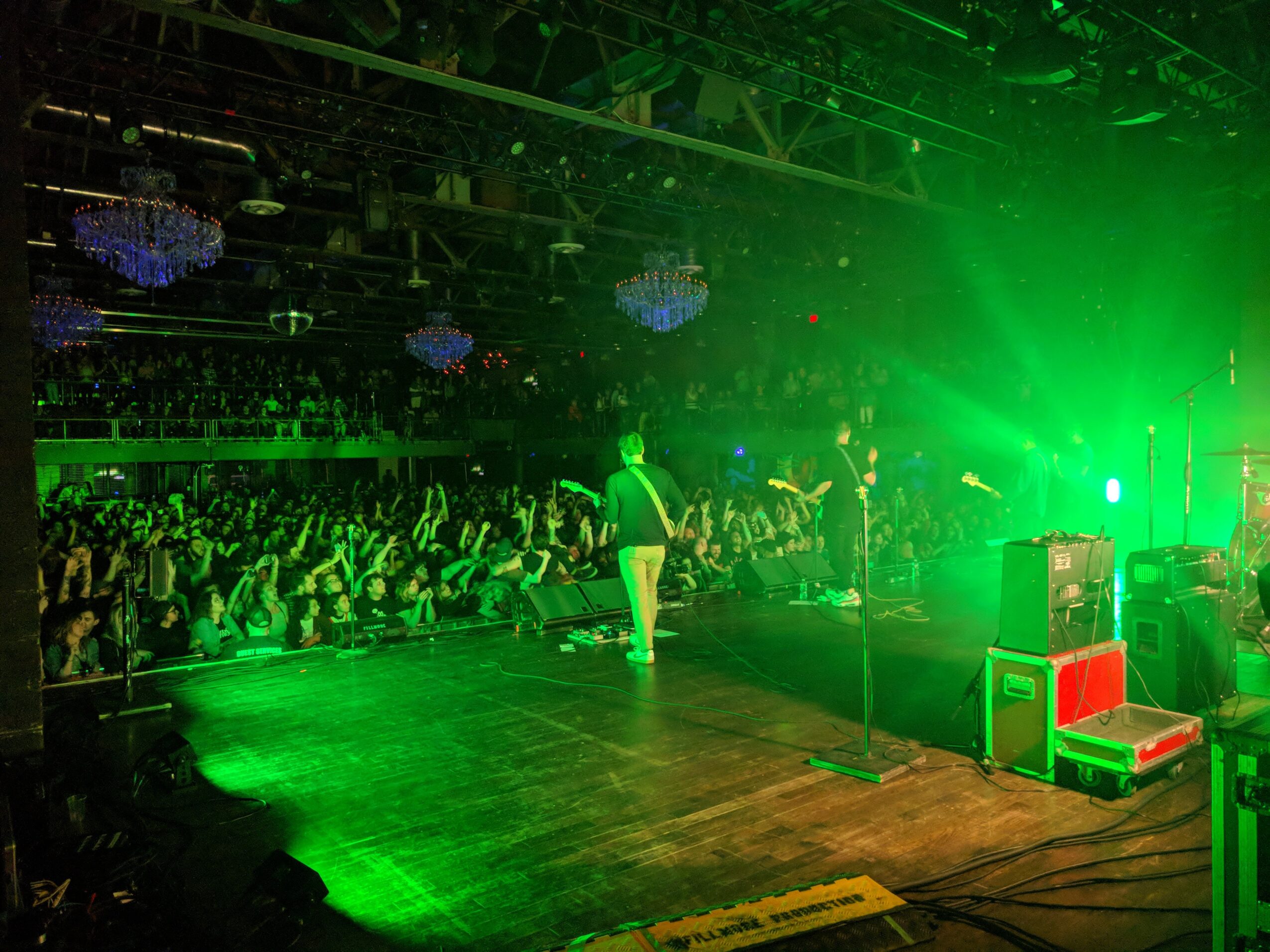 Performer on stage with green lighting, audience watching in a large indoor venue, stage equipment visible.