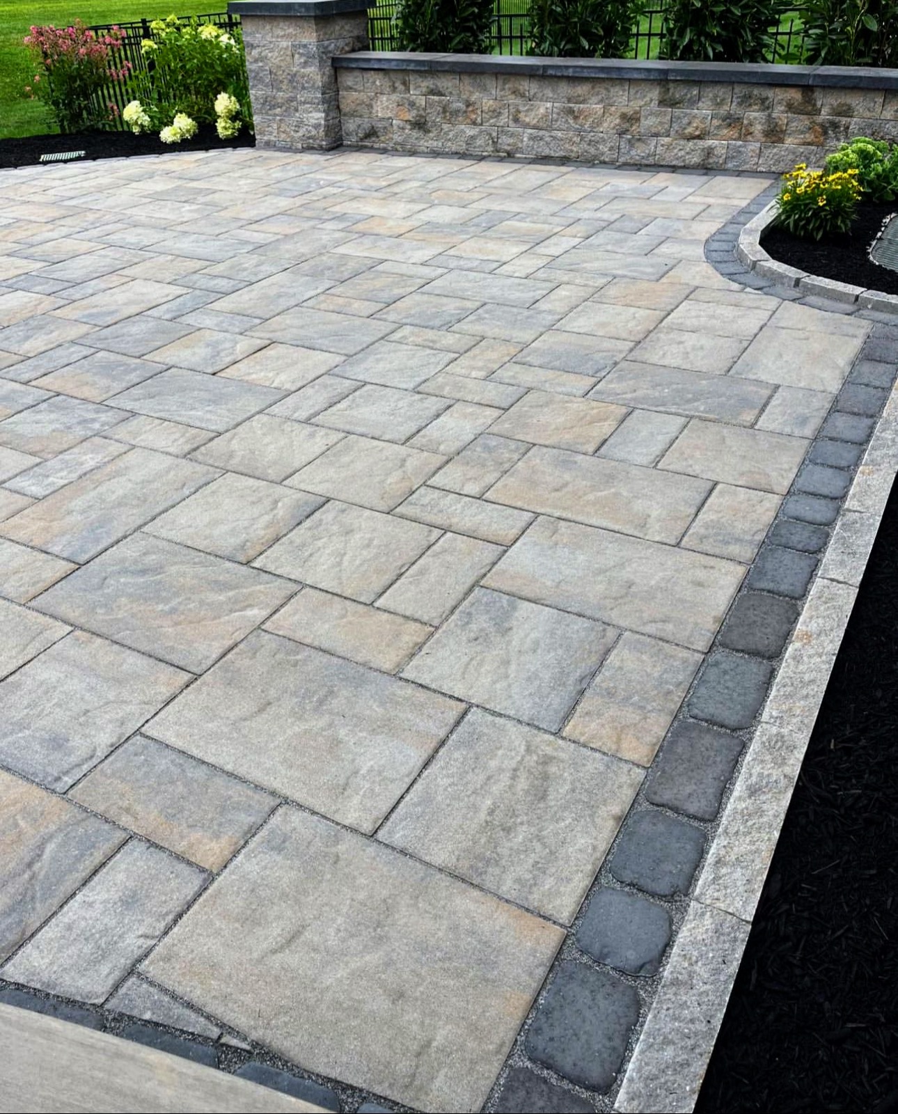 Paved outdoor area with stone tiles, bordered by darker stones and a garden with plants and flowers.