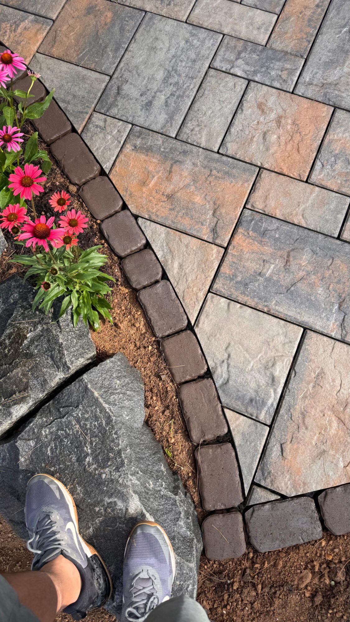 Footwear visible near a garden bed with pink flowers, bordered by dark bricks and paving stones.