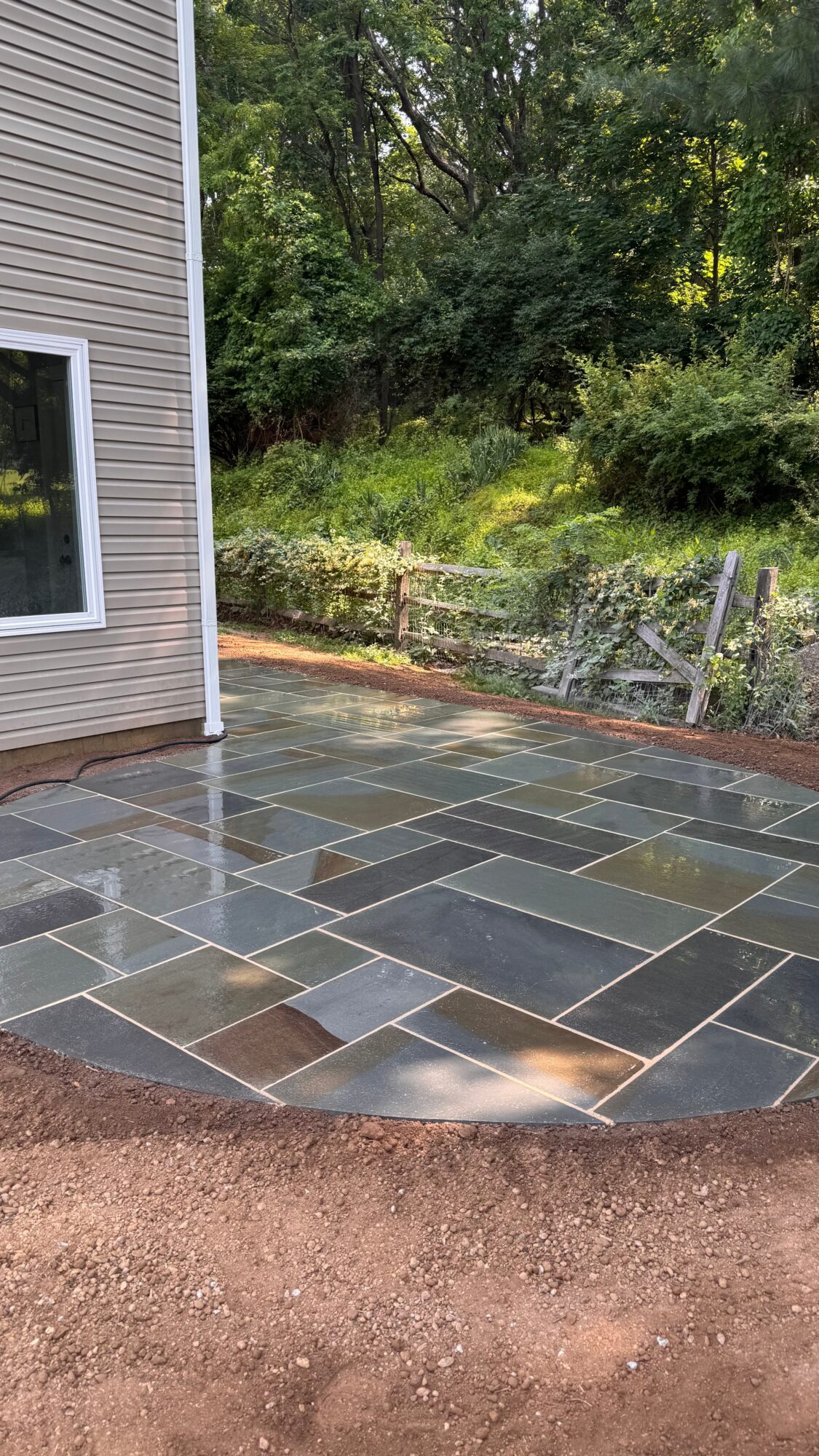 Newly paved stone patio next to house with siding, surrounded by soil and a wooded background.