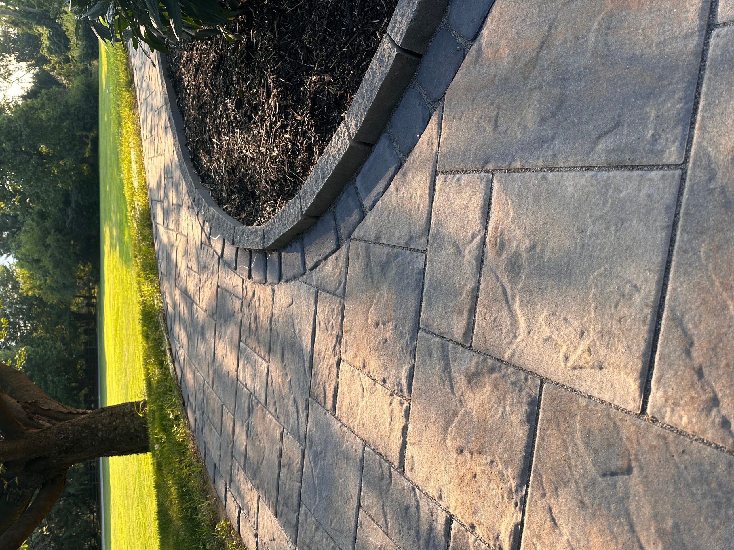 Curved stone pathway with a garden bed and grass on the side, viewed from an angle.