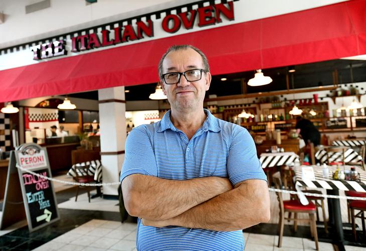 Man with glasses and a blue shirt stands with arms crossed outside a restaurant named The Italian Oven.