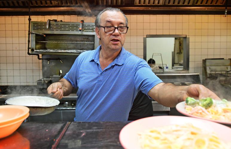 Man in blue shirt serving food in a kitchen with tiled walls and cooking equipment