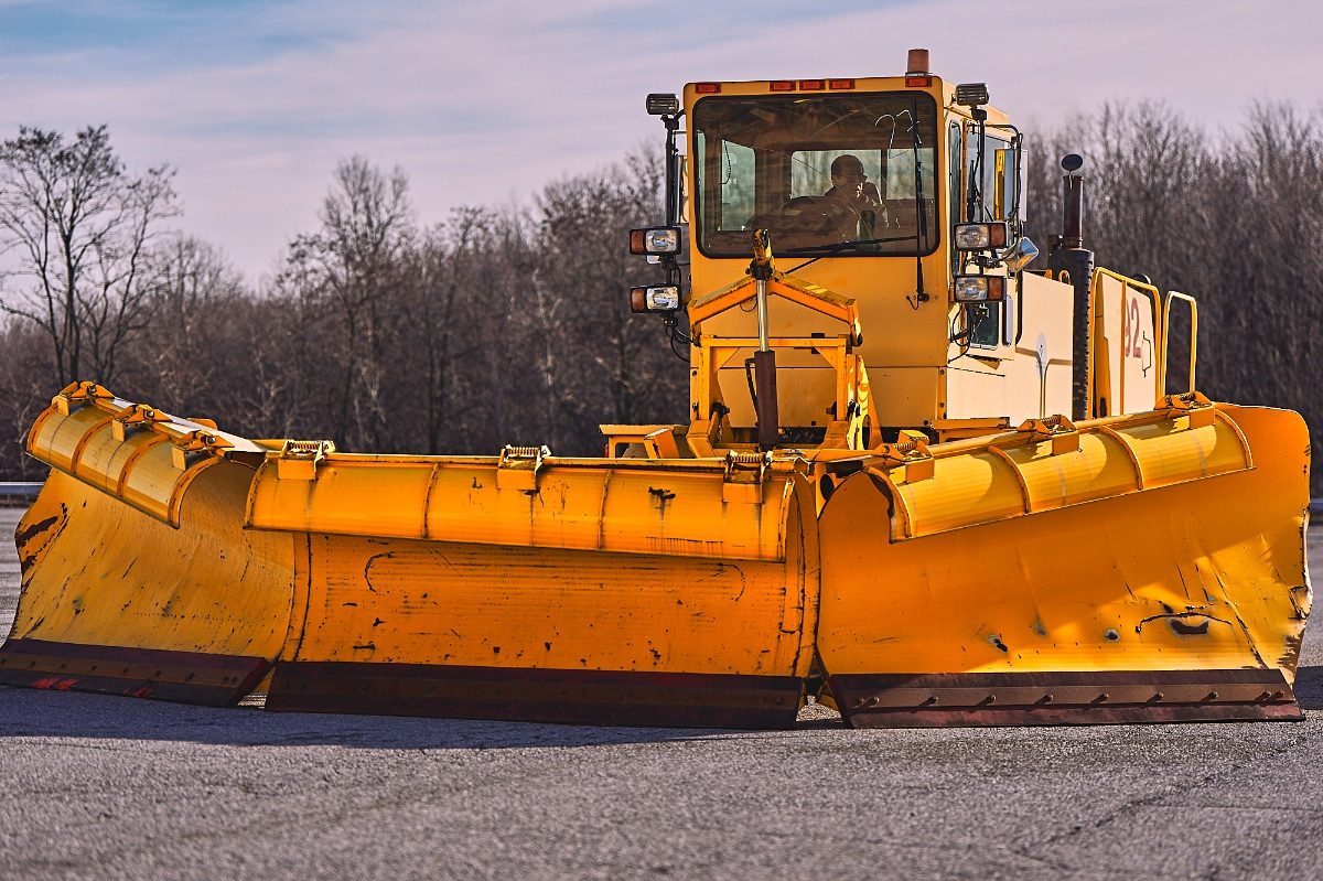 Front view of a large yellow snowplow with a wide blade, parked on a road with trees in the background.