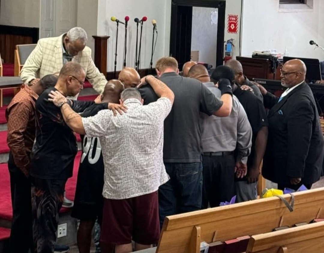 Group of people praying together with hands on each other's shoulders inside a church, with a man leading prayer.