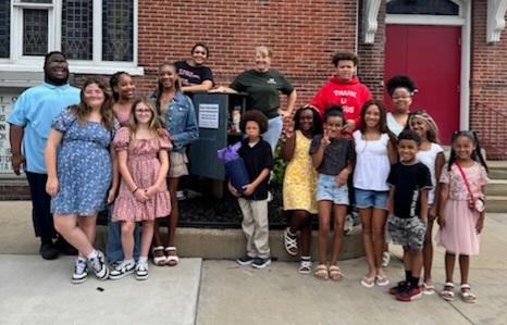 Group of children and adults standing outside a brick building, smiling, with some holding backpacks.