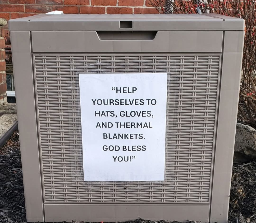 A gray outdoor utility box with a printed sign attached, containing a humorous message about hats, gloves, and blankets.